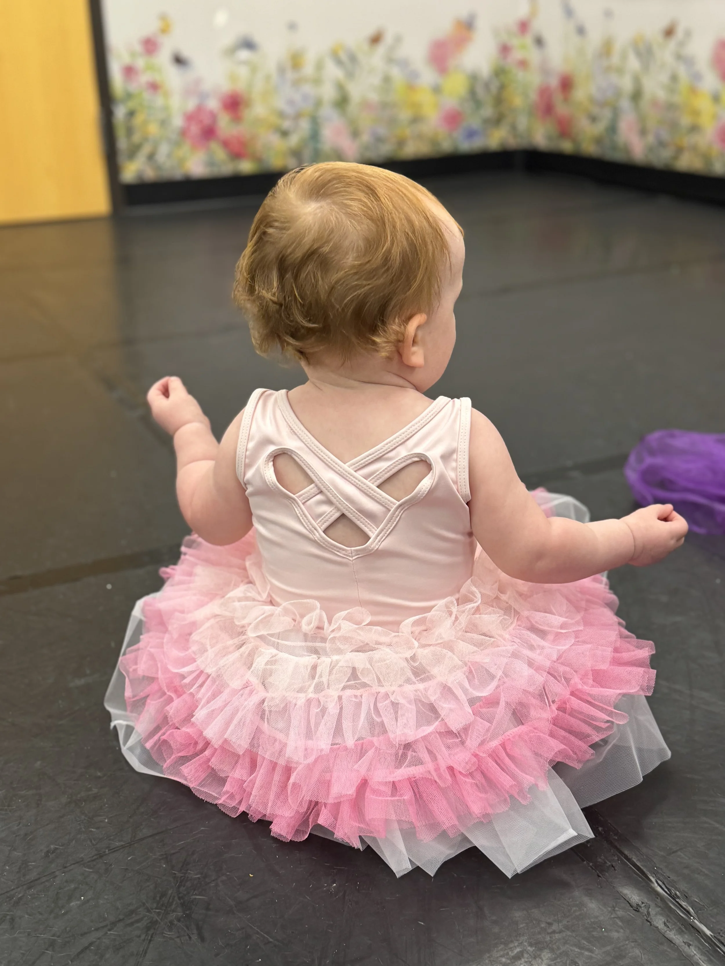 Back view of a baby girl wearing a pink tutu dress sitting on a black floor.