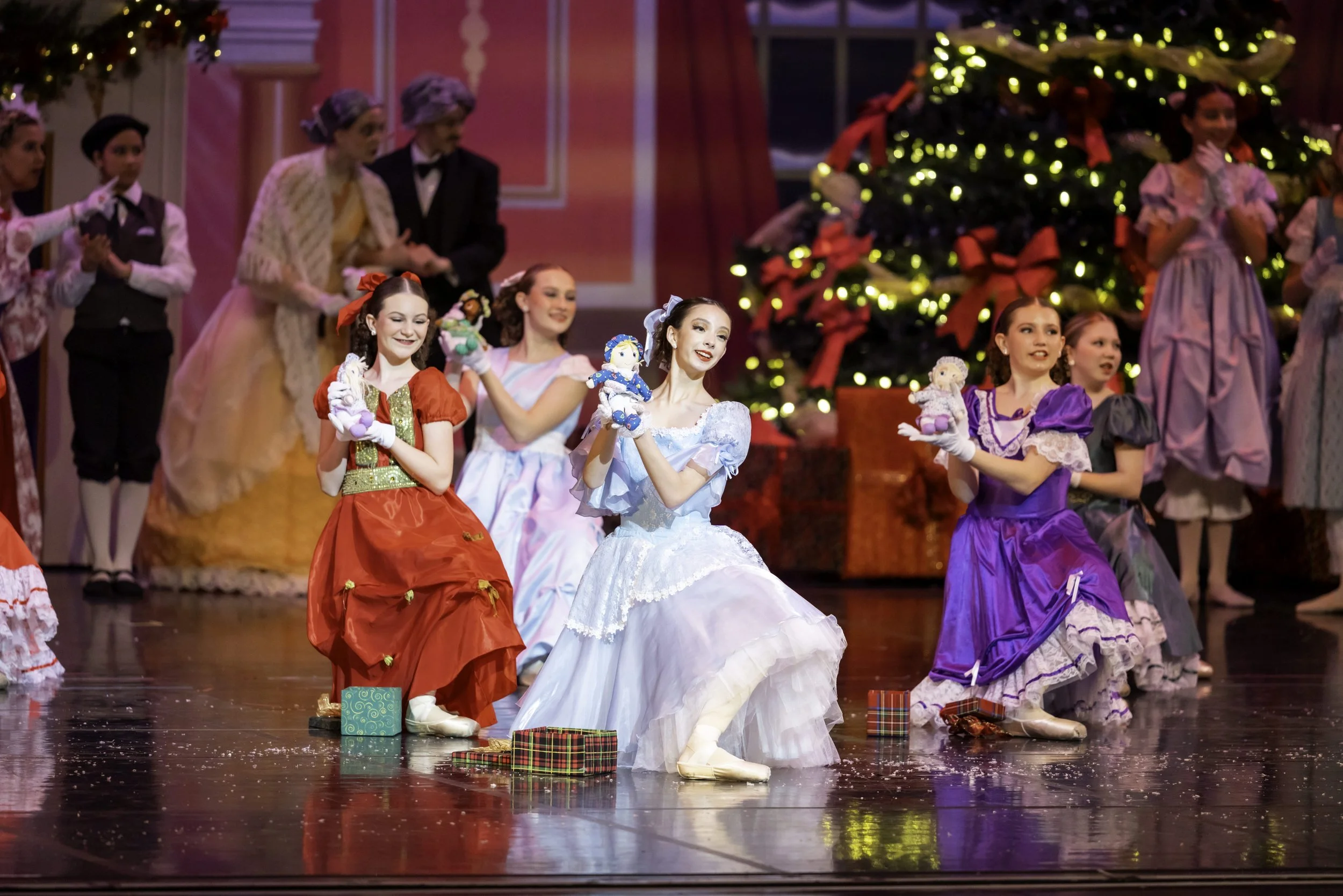 A group of children dressed in colorful costumes performing in a holiday-themed stage play with a Christmas tree and gifts in the background.