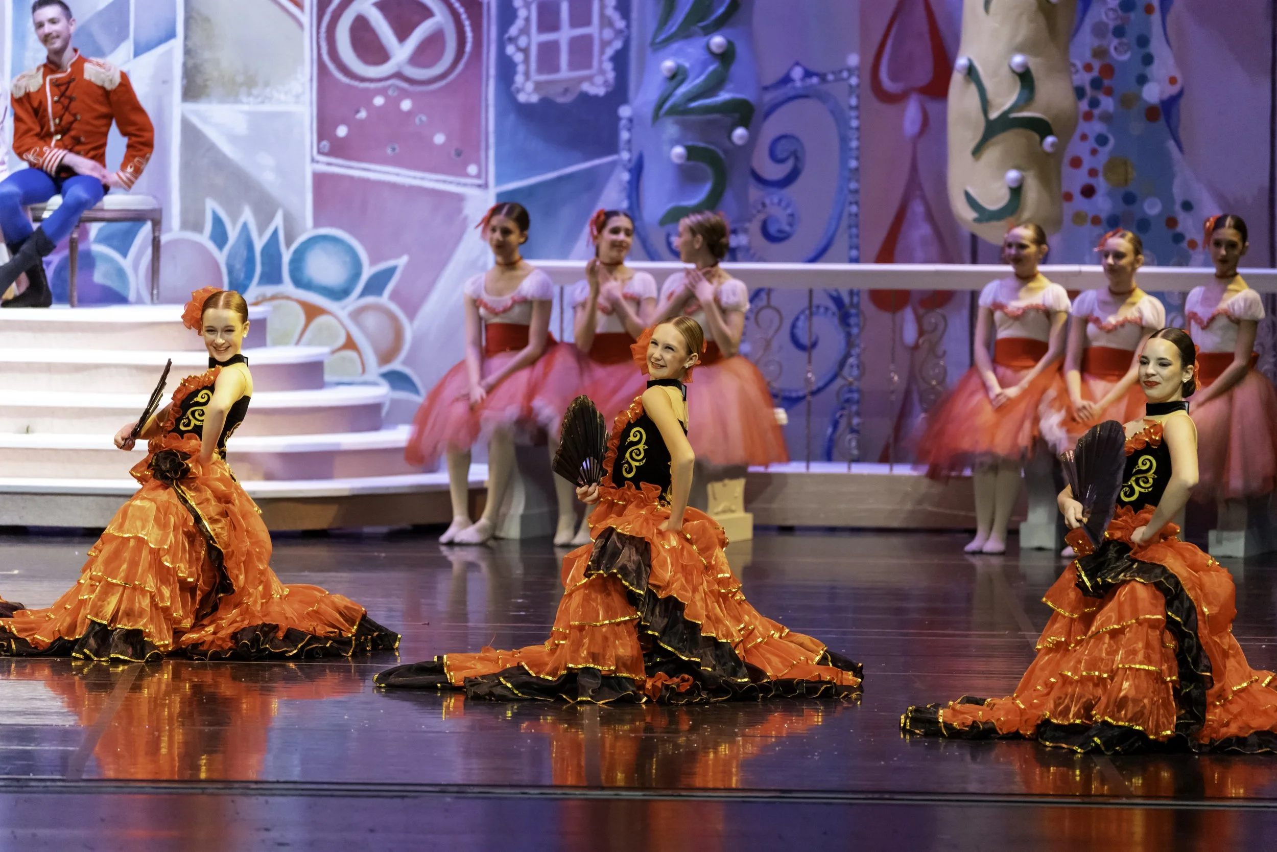 Girls in colorful costumes performing a dance on stage in front of a playful, decorated background with pink, blue, and yellow elements.