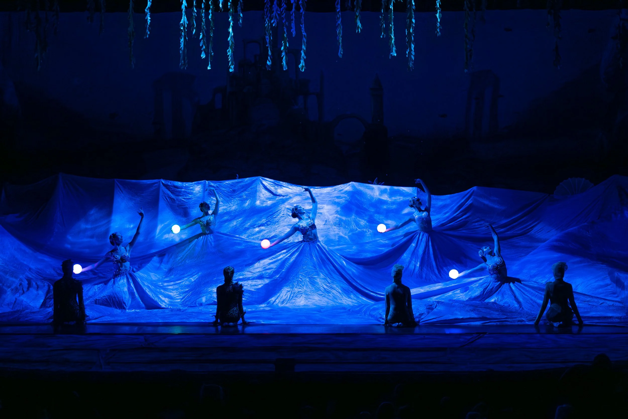 Ballet dancers performing on stage in a blue-lit scene with costumes and props simulating a night scene or underwater setting.