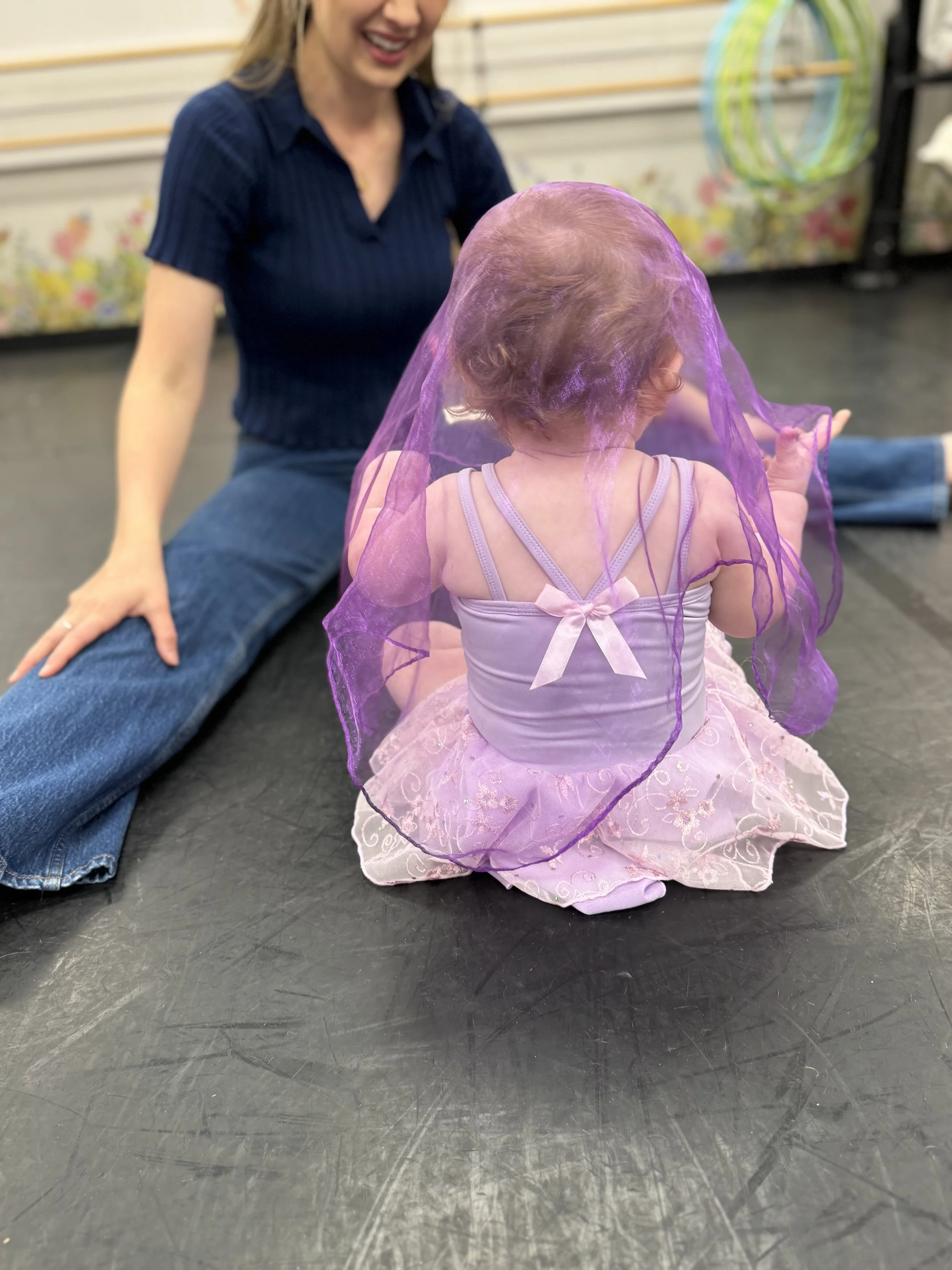 A young girl dressed as a princess with a purple veil and pink dress sits on the floor facing away, with a woman in a blue top and jeans sitting nearby, in a playroom or classroom setting.