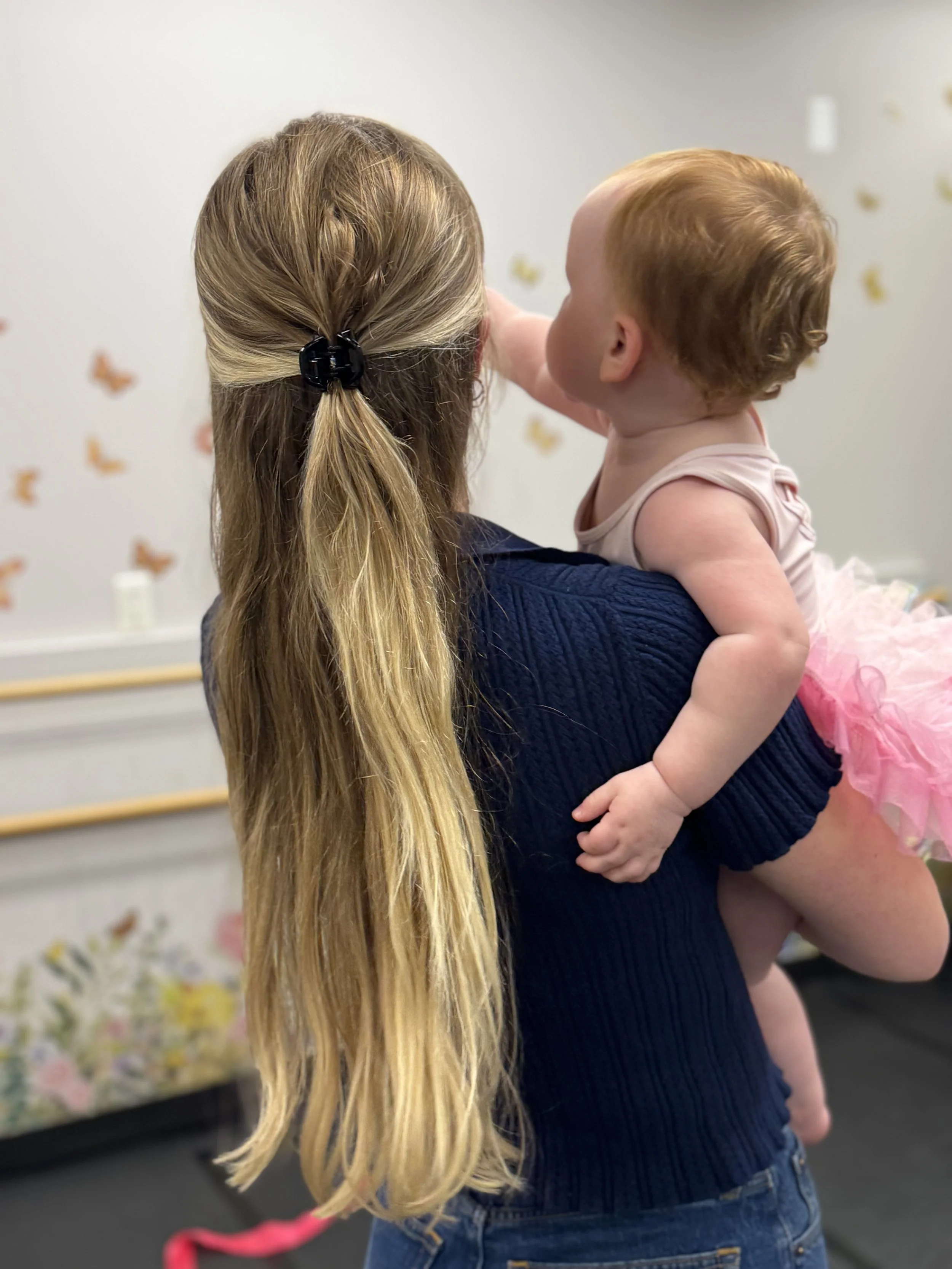 A woman with long blonde hair in a ponytail held with a black hair clip holds a toddler girl with red hair and light skin in her arms. The girl is wearing a pink tutu dress and reaching towards the wall decorated with butterfly stickers.
