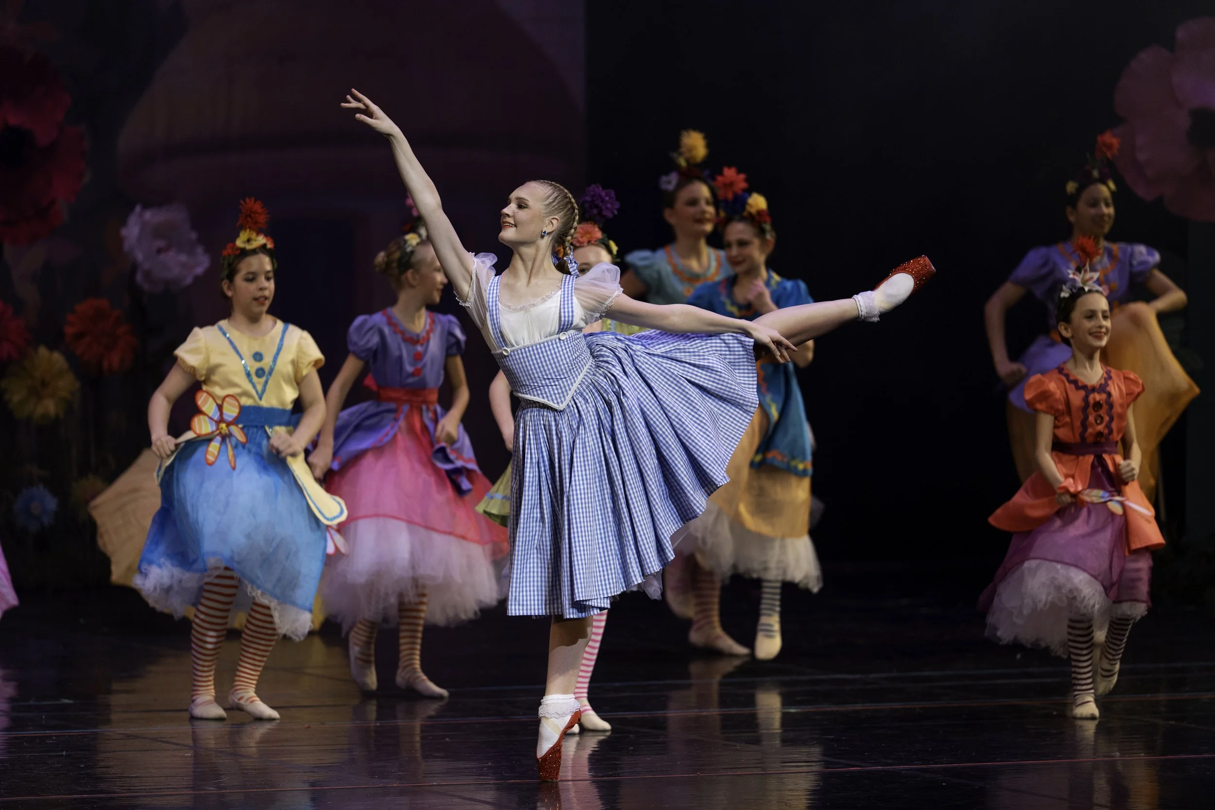 Young ballerinas in colorful costumes performing on stage with floral decorations in the background.