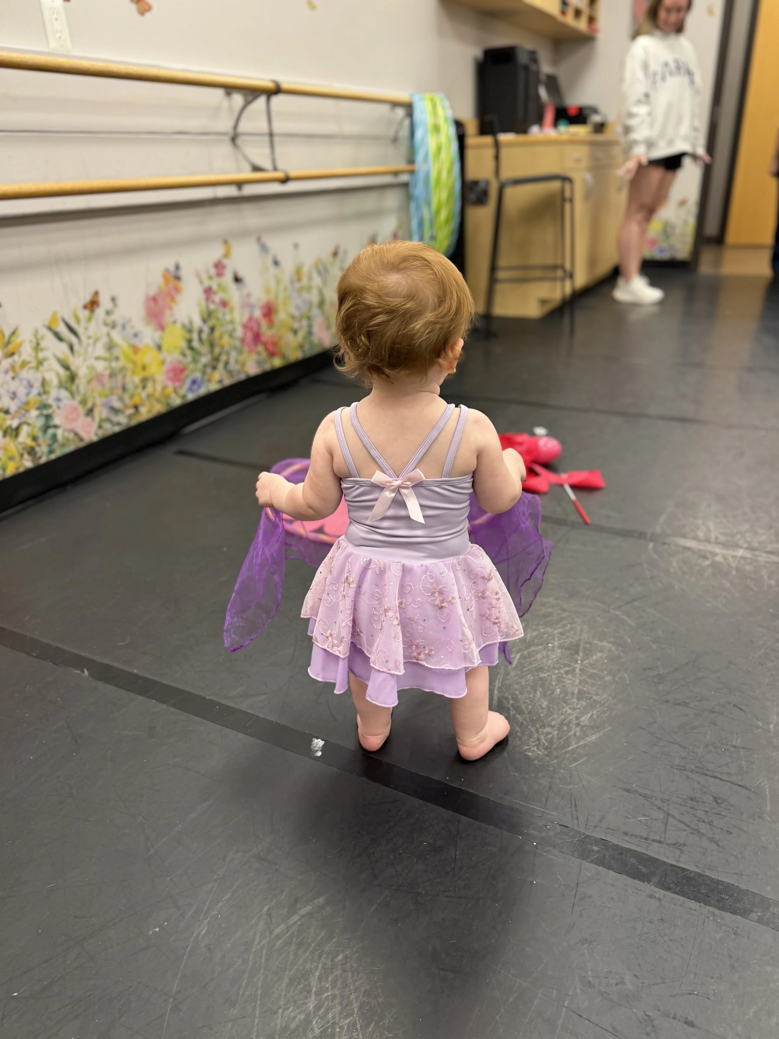 A young child in a purple fairy costume with a tutu and wings is standing barefoot on a dance studio floor, facing away from the camera.