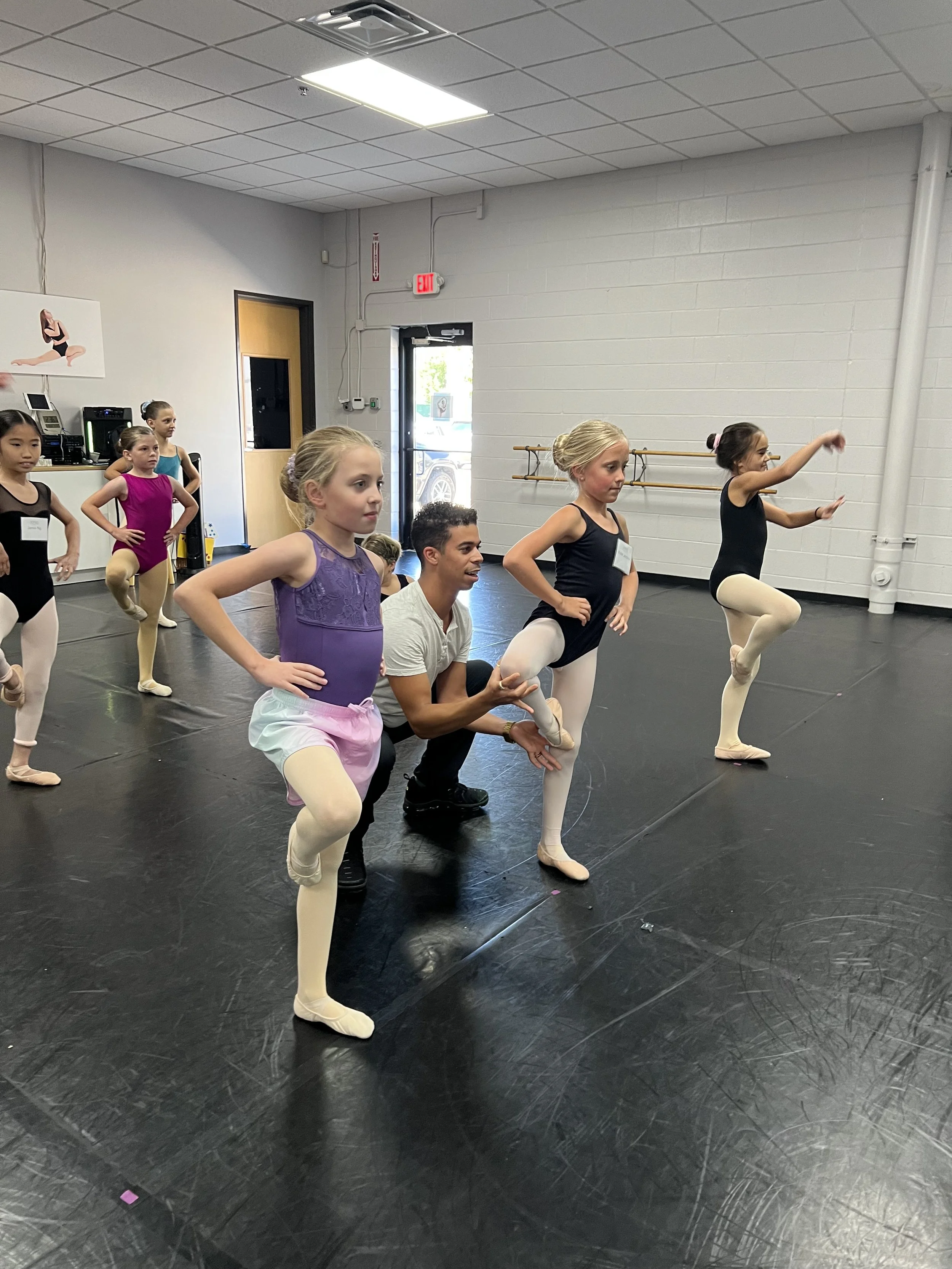 A ballet class with young girls practicing dance moves, supervised by an instructor, in a dance studio with mirrors, ballet barres, and a black floor.