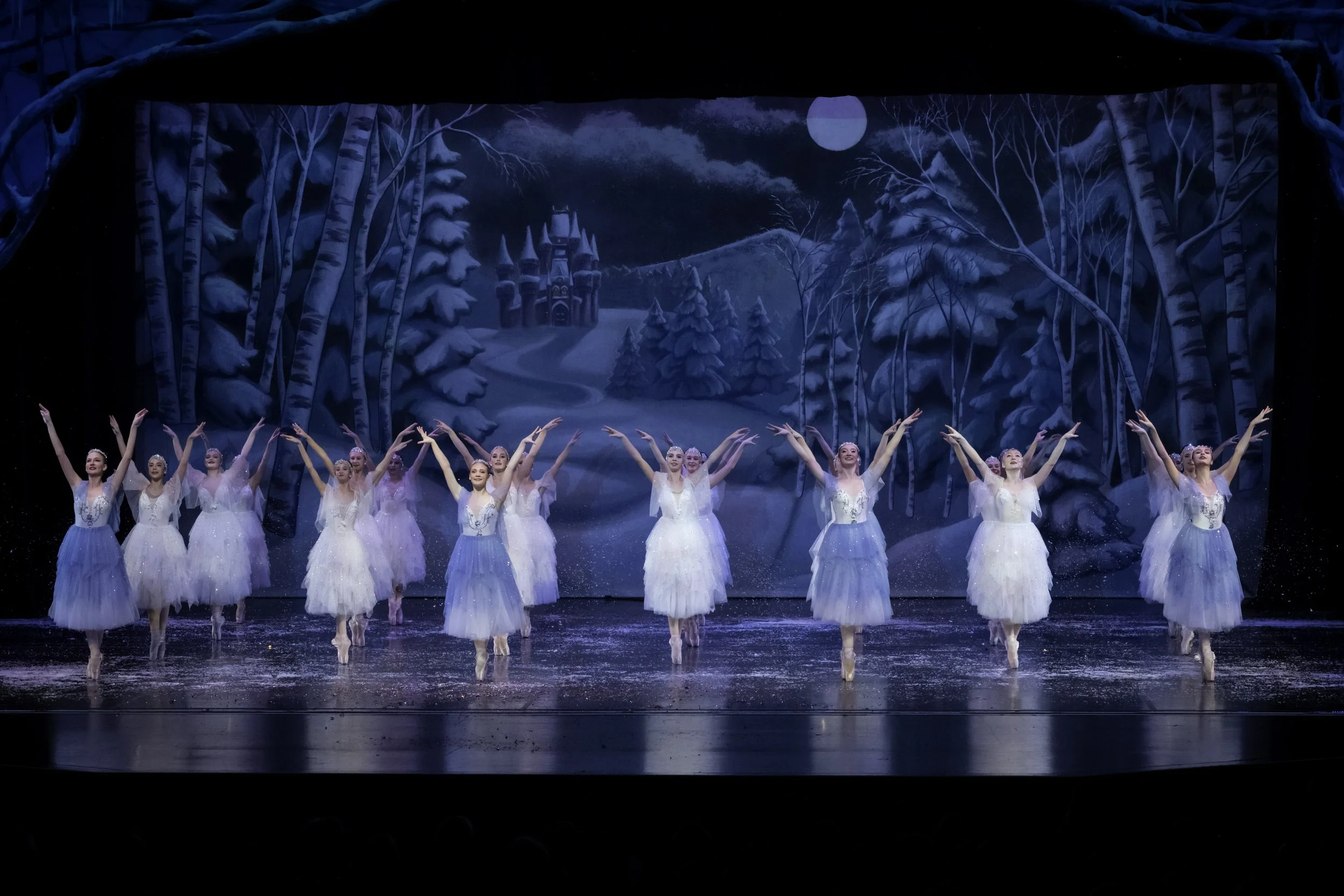 Ballet dancers in white and blue tutus perform on a stage with a winter landscape backdrop, featuring snow-covered trees, a castle, and a moon.