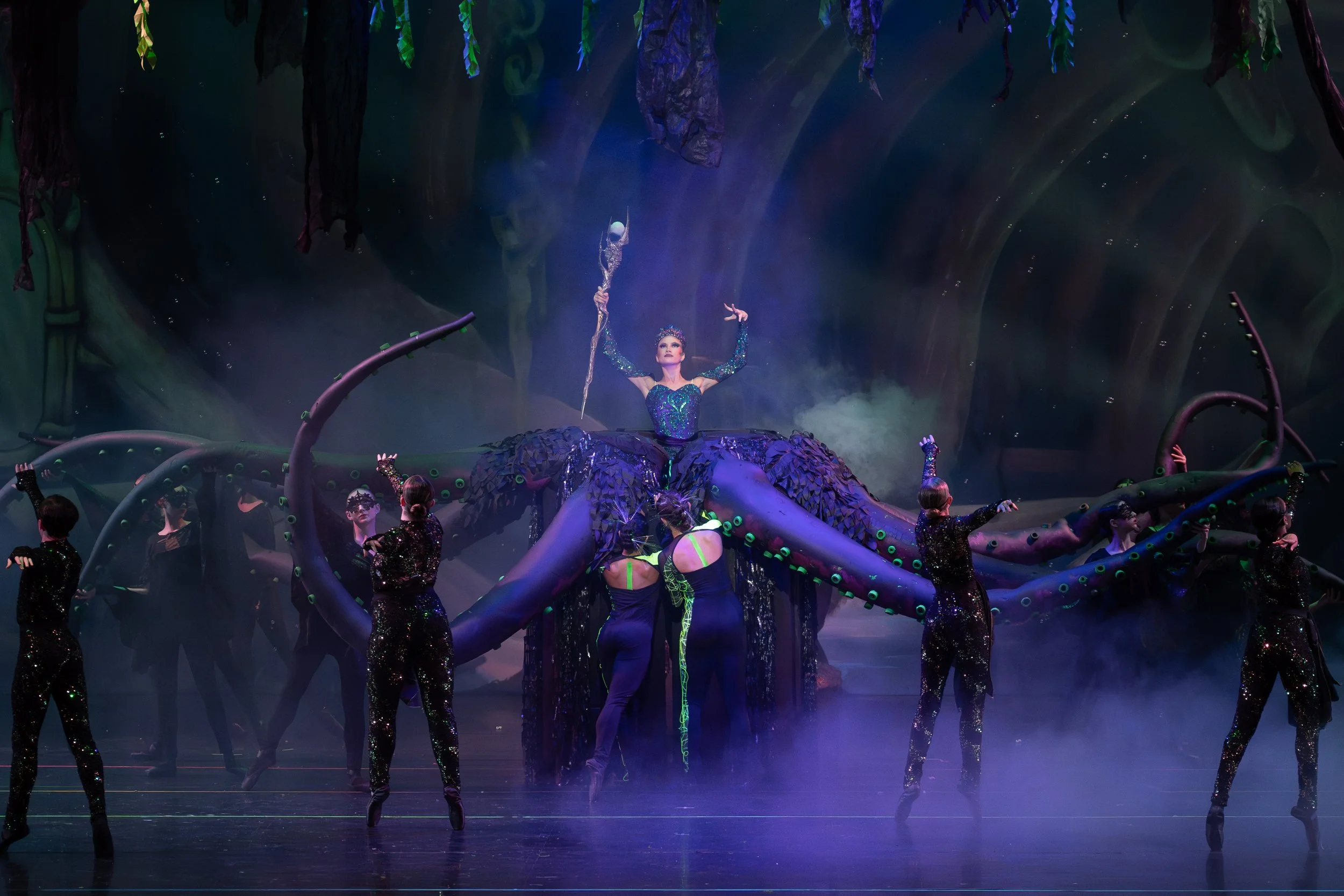 Performer dressed as a grapevine queen with a large purple gown and crown, holding a staff, surrounded by dancers in black costumes, on a stage with a forest backdrop and misty atmosphere.