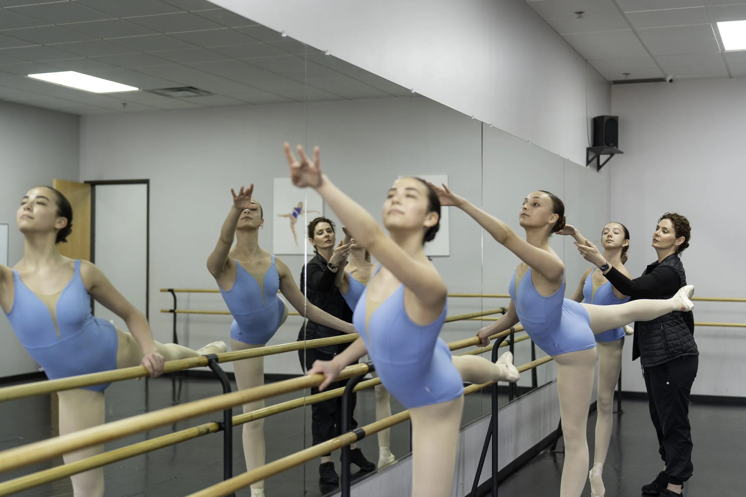 Ballet dancers in blue leotards practicing at a ballet barre with instructors in a dance studio.