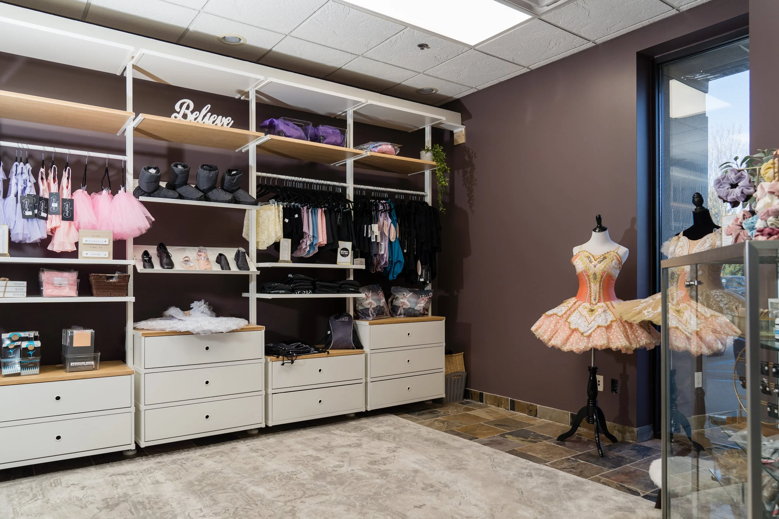 Dance costume shop with a ballet dress on a mannequin, ballet slippers, tutus, and dancewear on shelves and racks.