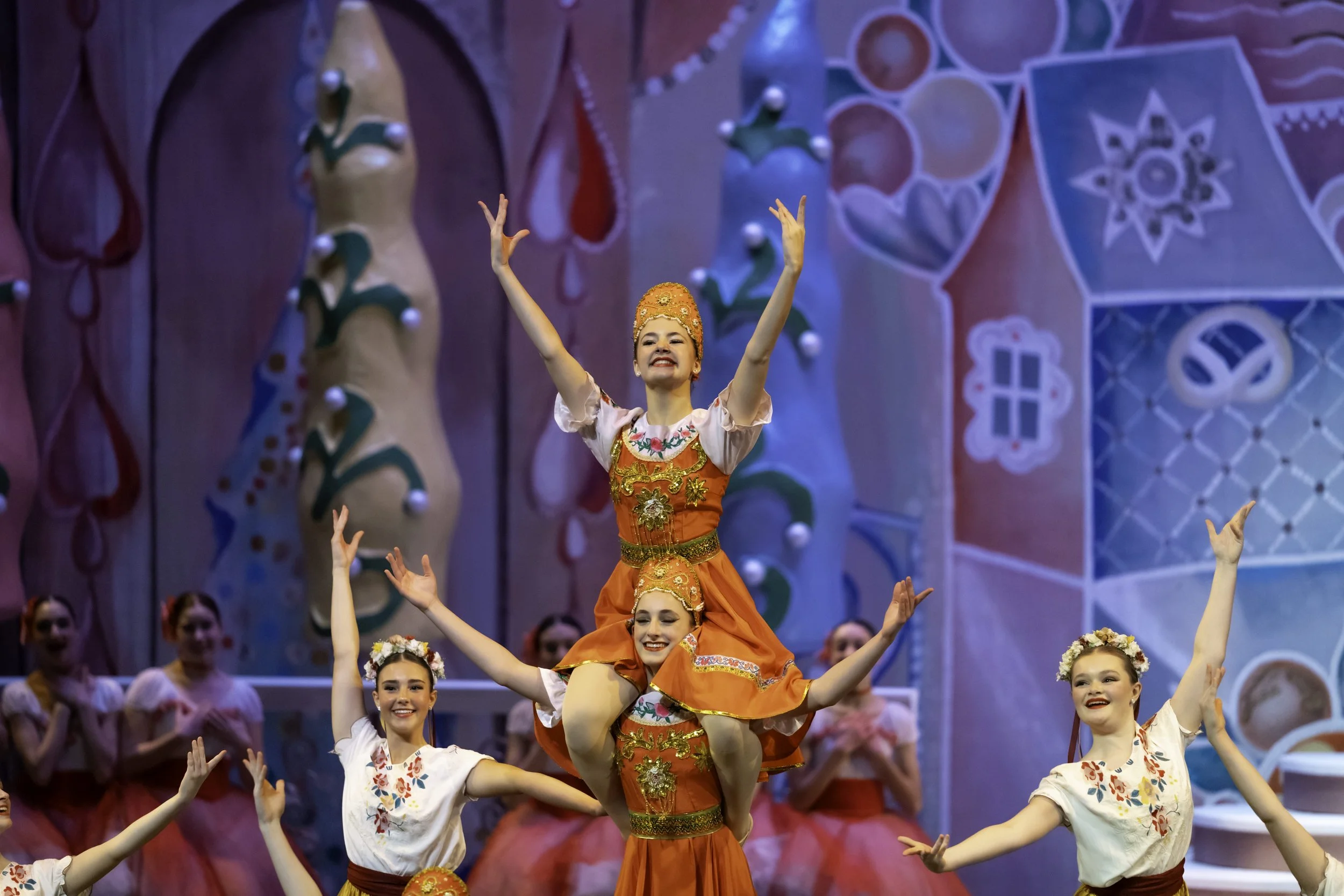 Performers in colorful costumes dancing on stage with a backdrop resembling a gingerbread house.