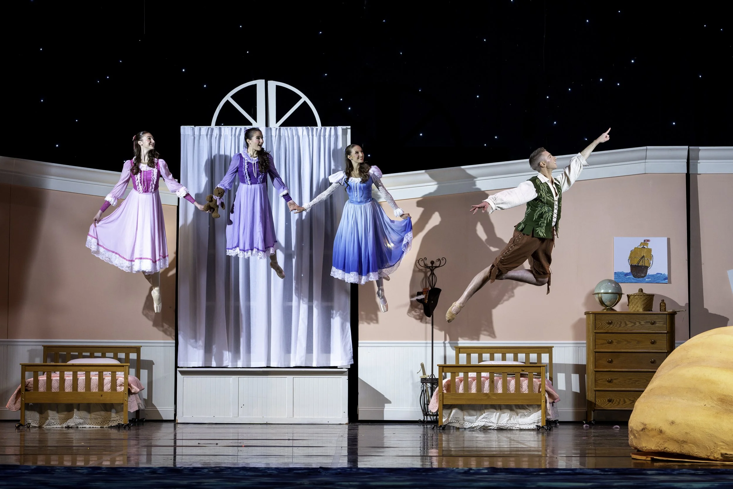 Stage scene from a theatrical production with four women in colorful dresses and one man in historical costume, all in mid-air jumps, holding hands or reaching out, with a bedroom set and starry night backdrop.