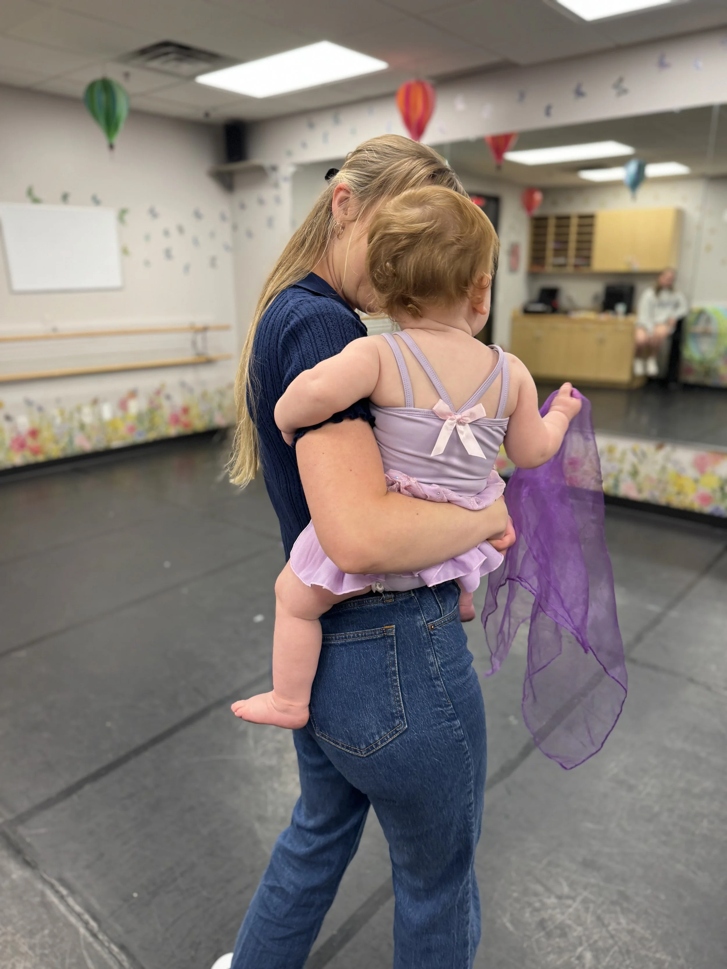 A woman holding a young girl indoors in a decorated room. The woman has long blonde hair and is wearing a blue shirt and jeans. The girl has light brown hair and is wearing a purple dress with a pink bow and a purple cape. The background features han