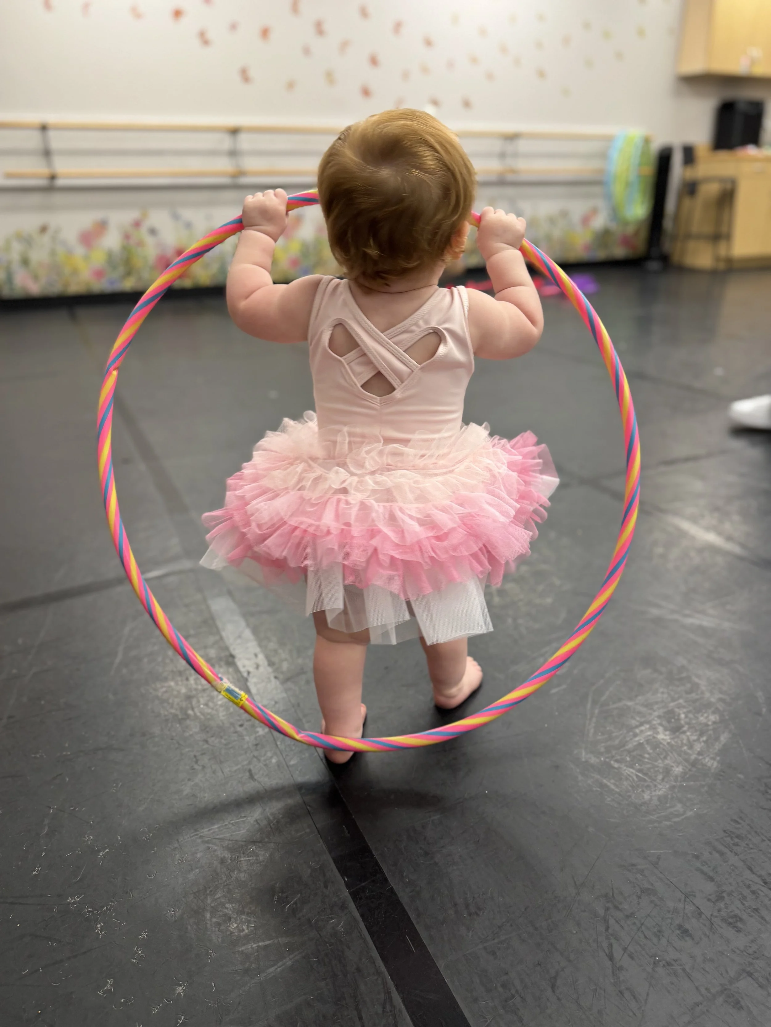 A young child with curly red hair wearing a pink tutu dress and white tights, holding a colorful hula hoop while standing on a black dance studio floor.
