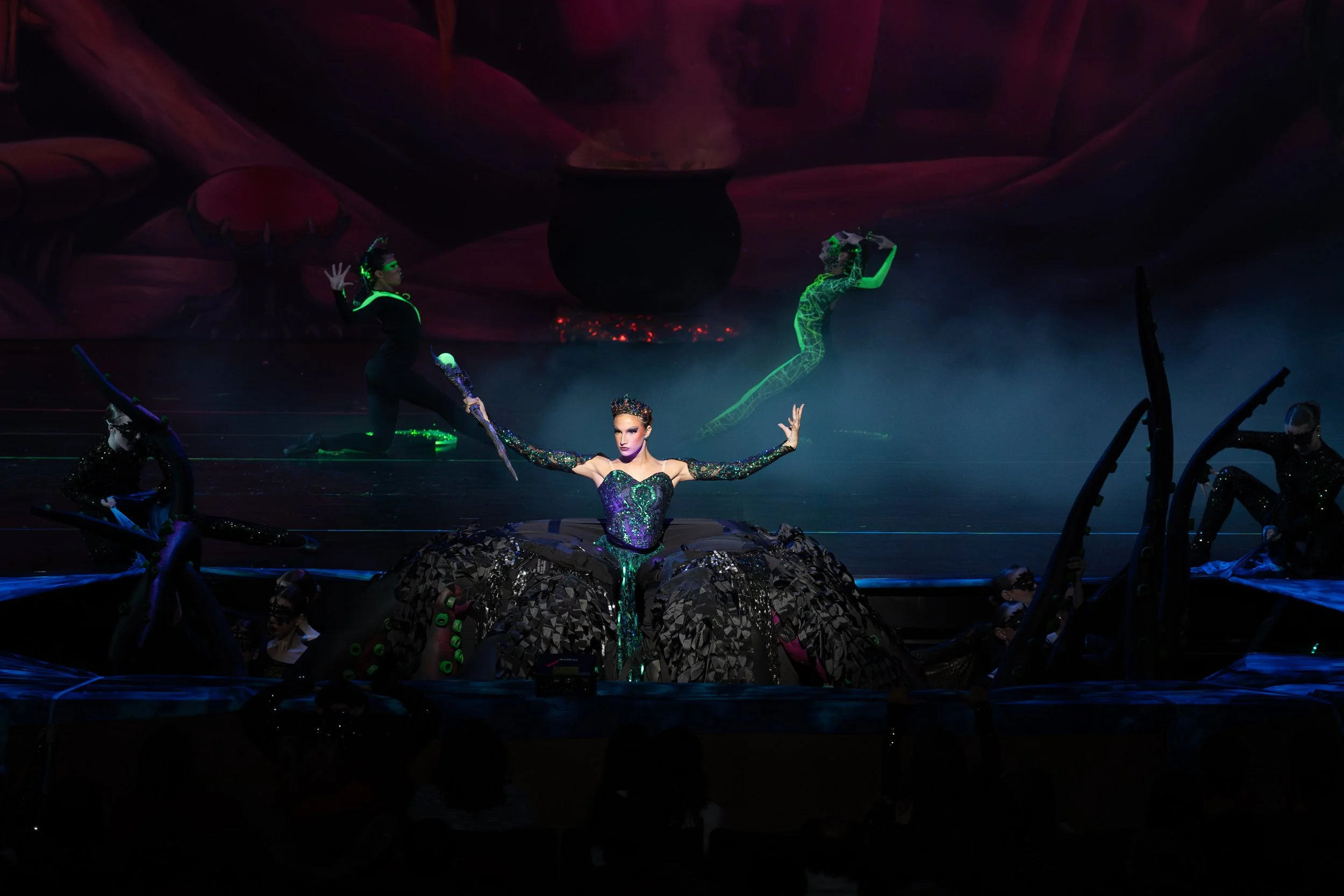 A theatrical stage performance featuring a central female performer dressed in an elaborate black and purple costume with large, decorative skirt, surrounded by dancers in black costumes with glowing green and blue accents, set against a dark, dramat