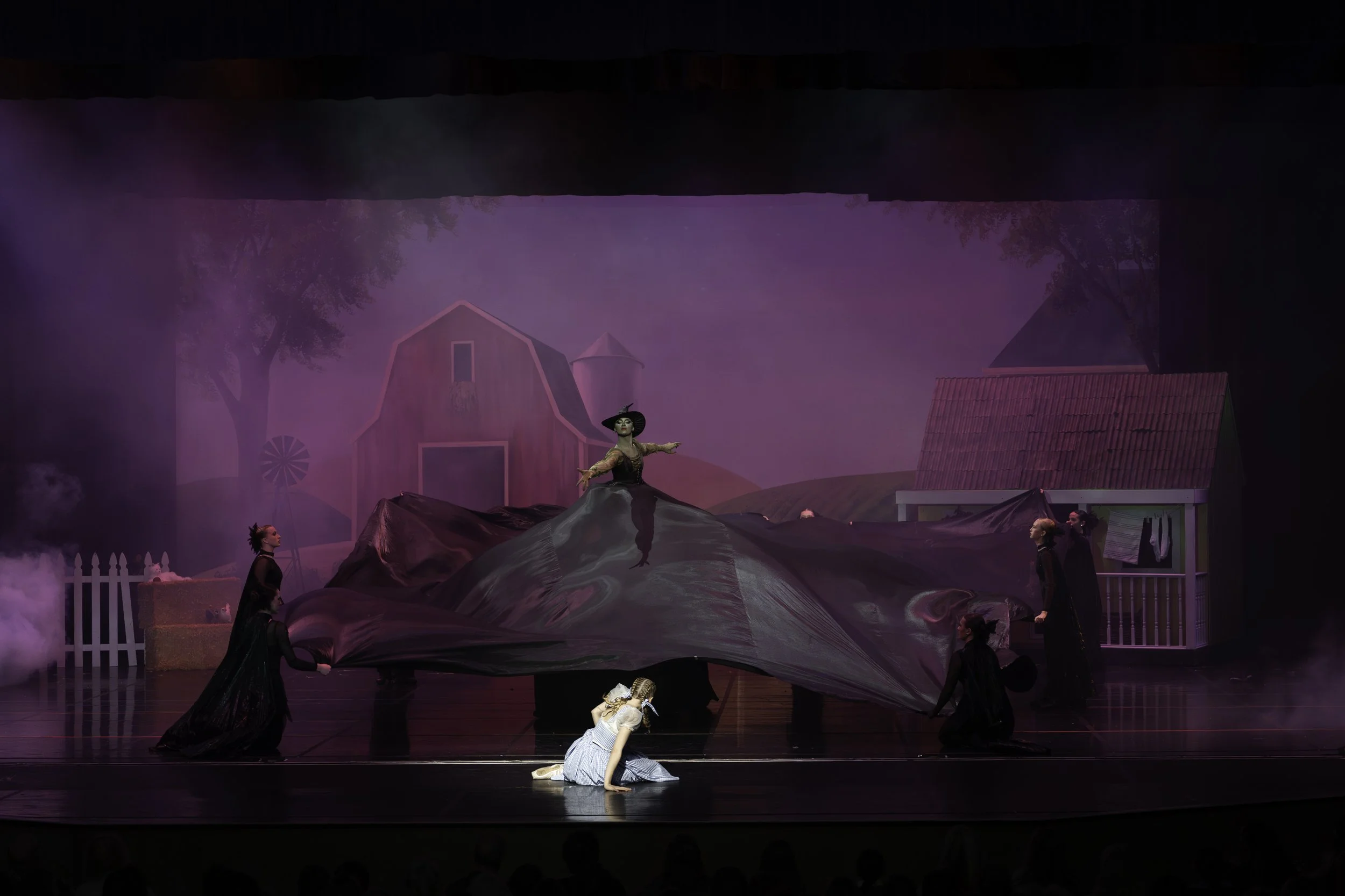 Theatrical stage scene depicting characters in a farmyard with a large black fabric, a girl in a blue dress kneeling on the floor, a woman in costume standing on a mound of fabric, and other performers around. Background includes a barn, a small hous