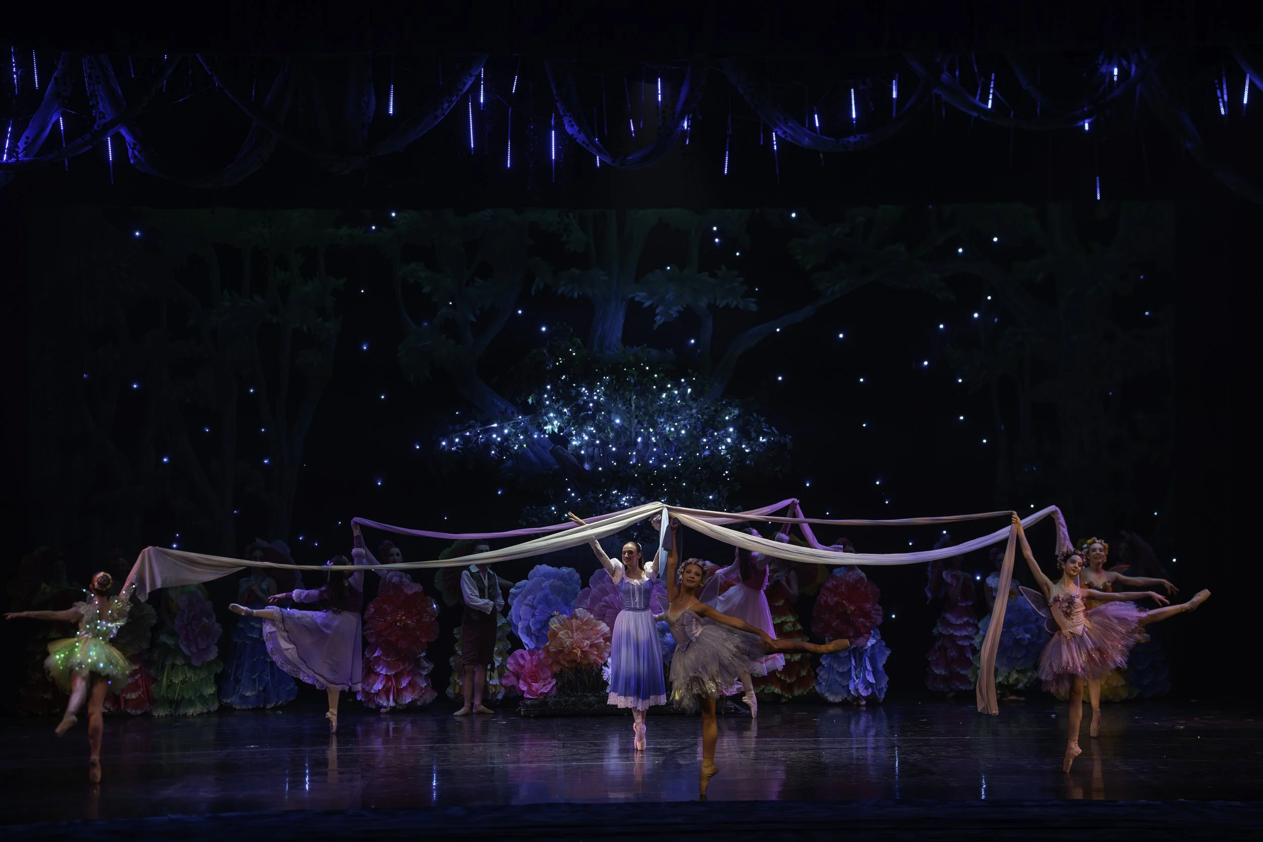 Ballet dancers perform on stage with a backdrop of twinkling lights and a tree, surrounded by colorful oversized flowers.