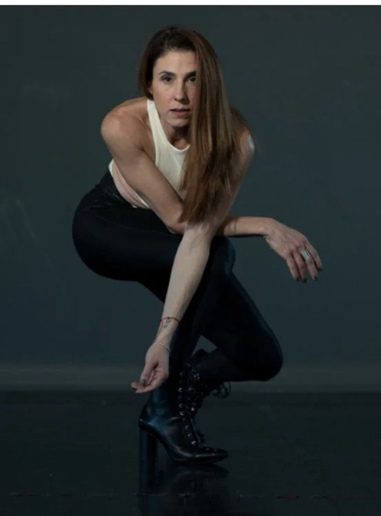 A woman with long brown hair, wearing a white sleeveless top, black pants, and black high-heeled boots, crouching and looking directly at the camera against a dark background.