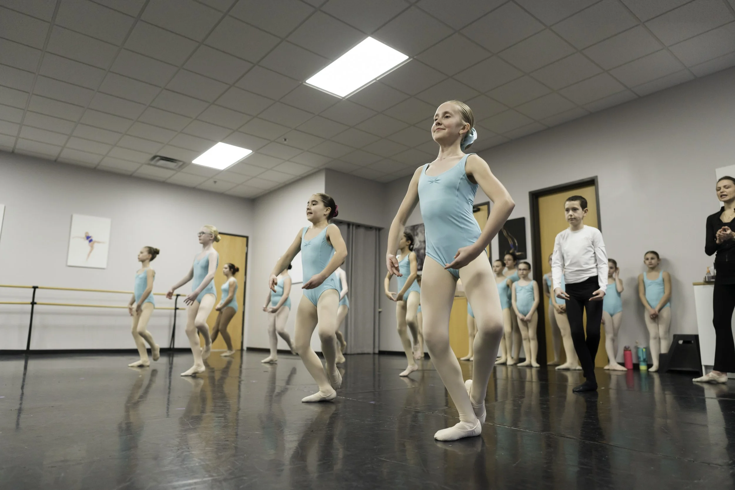 Group of young ballet dancers in blue leotards and white tights practicing in a dance studio, with their instructor observing.