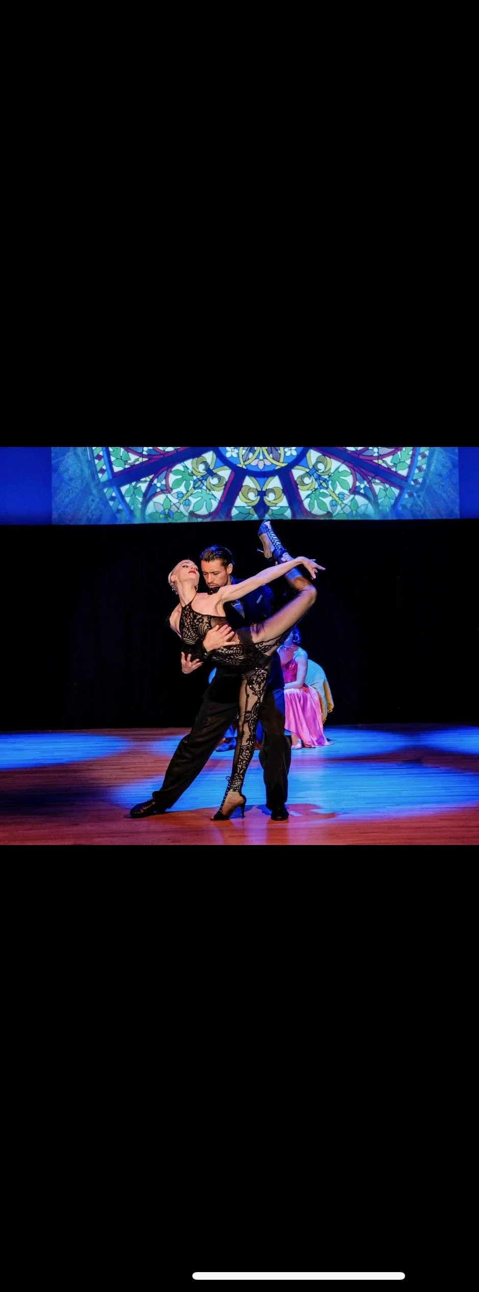 A male and female dance pair perform a passionate ballroom dance on stage, with a stained glass window projected in the background and another person sitting in a pink dress in the background.