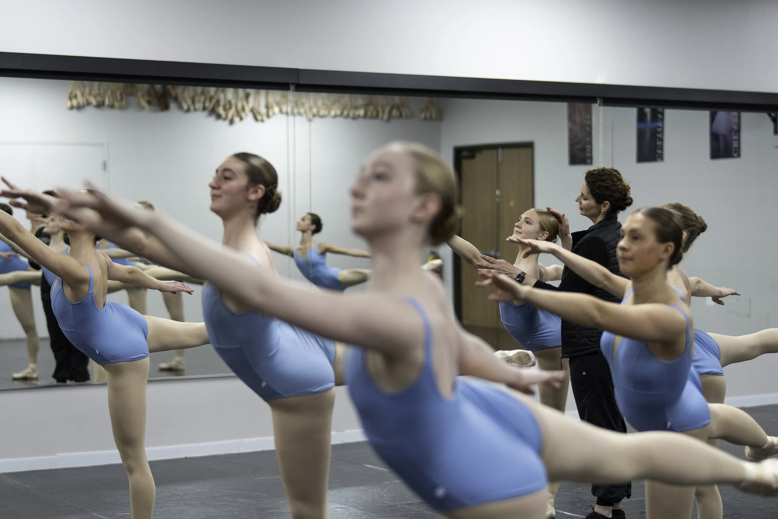 Ballet students practicing dance moves in a dance studio with mirrors, led by an instructor.