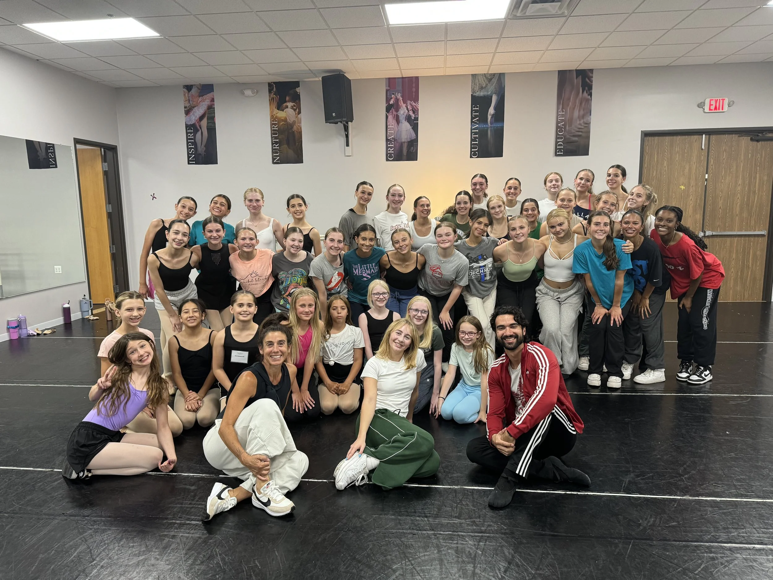 Group of young ballet dancers and two instructors posing in a dance studio with motivational banners on the wall.
