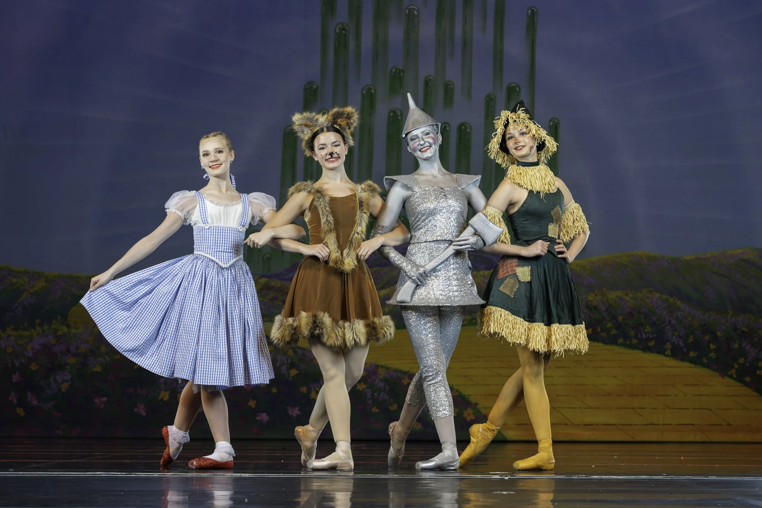 Four young girls dressed as characters from The Wizard of Oz performing on stage with painted backdrop of green towers and a yellow brick road.