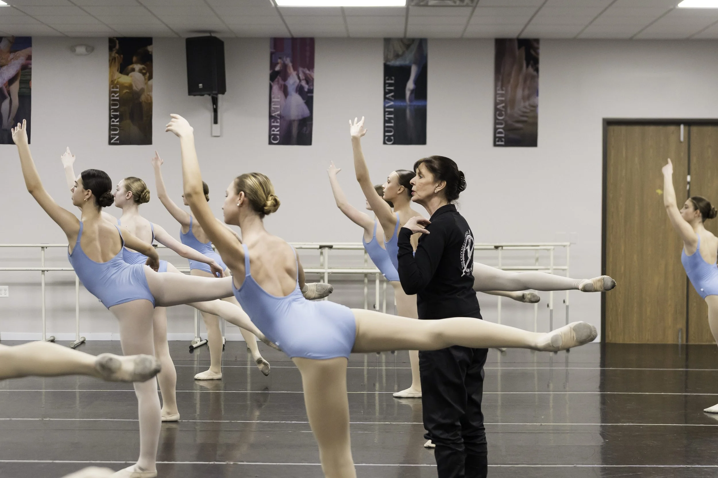 Ballet students practicing in a dance studio with an instructor observing, all wearing blue leotards and tights, with posters on the wall displaying dance-related words.