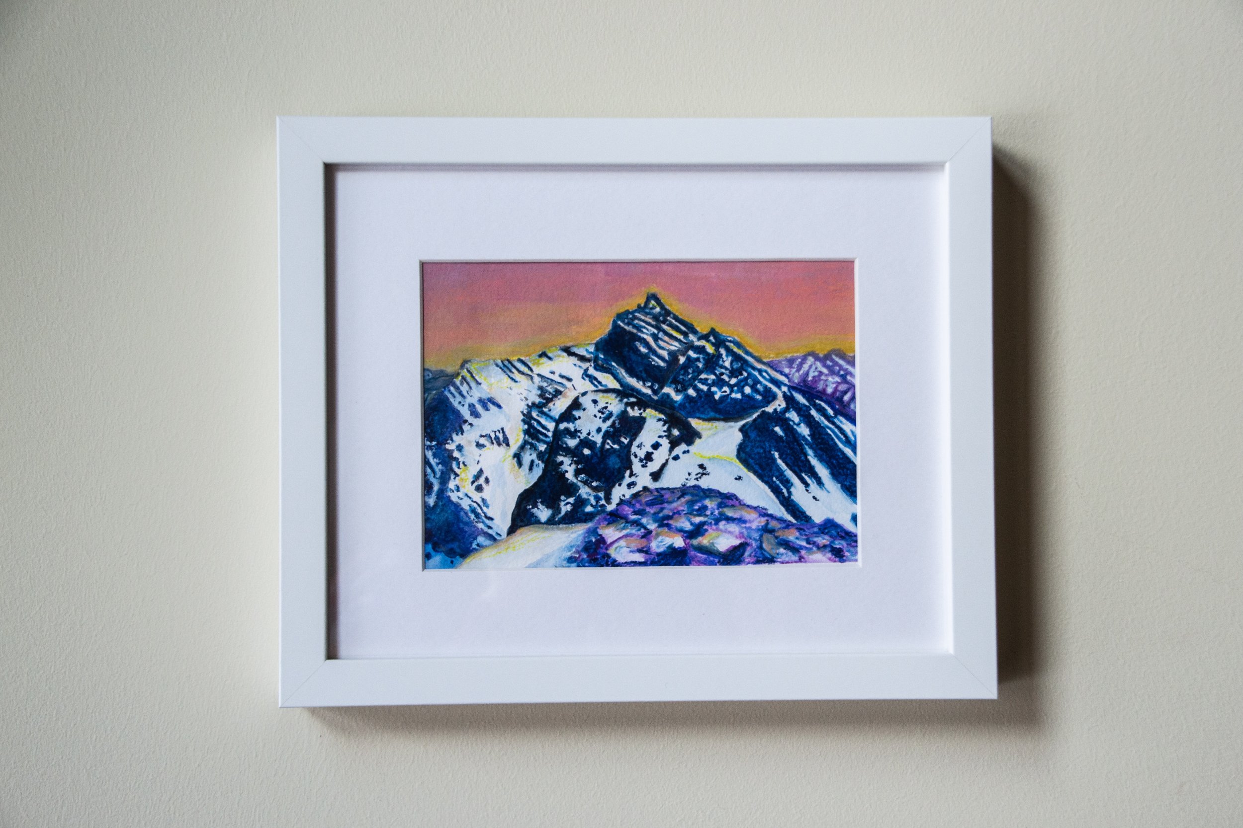 Framed painting of snowy mountain peaks at sunset with a colorful sky.