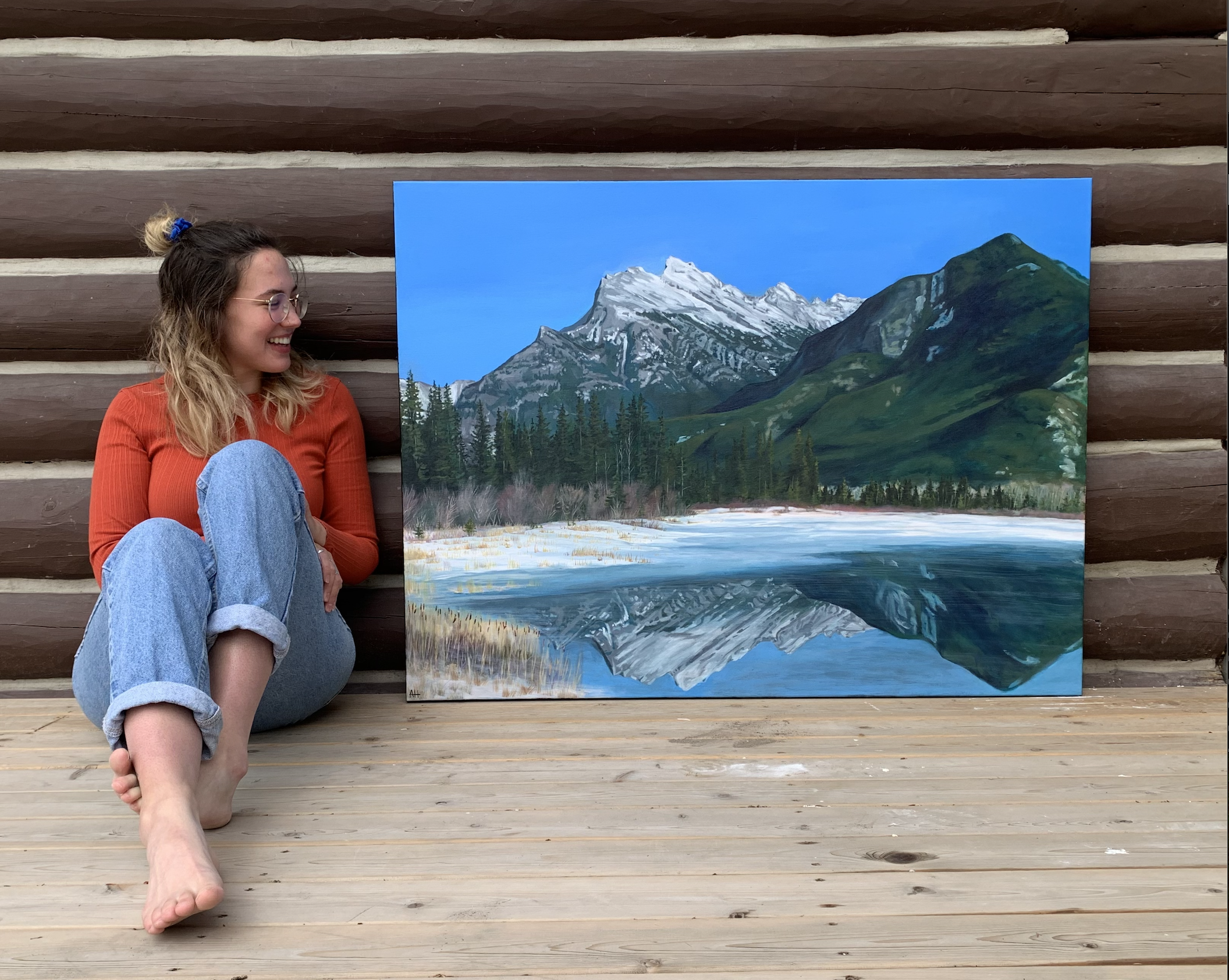Person sitting on wooden floor next to a landscape painting of a mountain and lake.