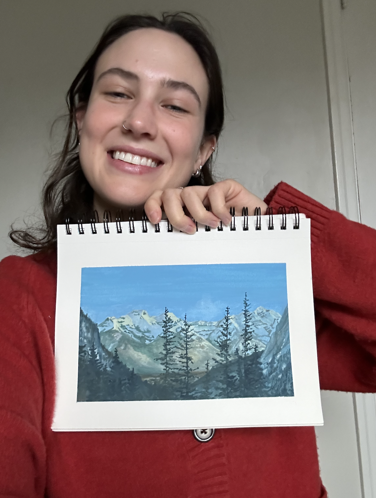Person smiling, holding a sketchbook with a painting of a mountain landscape.