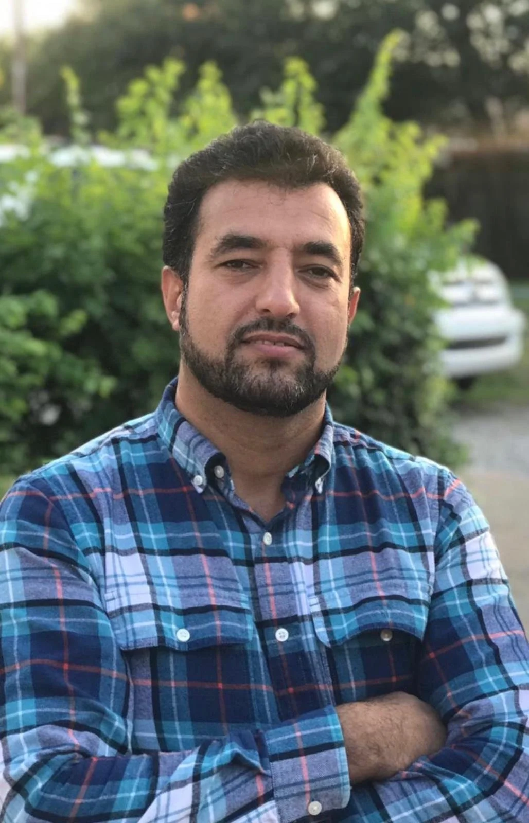 A man with dark hair and a beard wearing a blue plaid shirt, standing outdoors with greenery and a white car in the background.