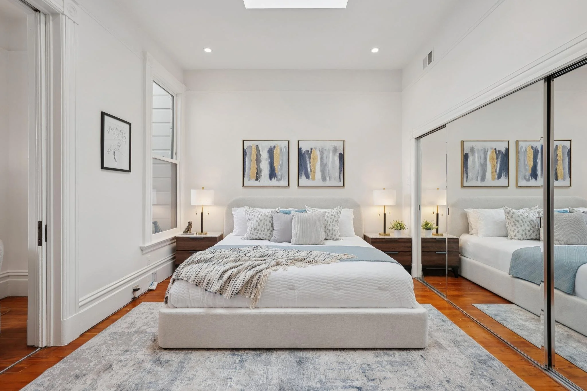 A modern bedroom with a white bed, two wooden nightstands with lamps, abstract wall art, and mirrored closet doors.