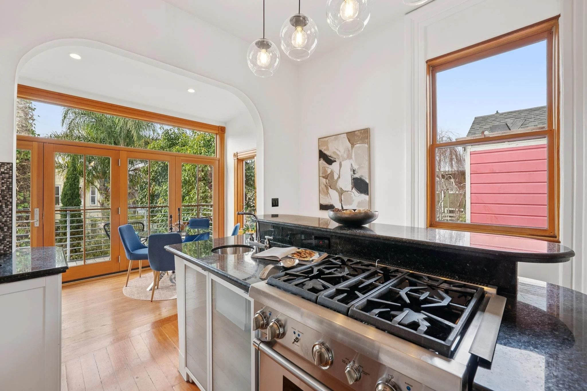 Kitchen with black granite countertops, stainless steel gas stove, white cabinetry, and a bar-height counter. There is a white wall with a abstract art piece and a window with wooden trim. A dining area with blue chairs and a small round rug is visib