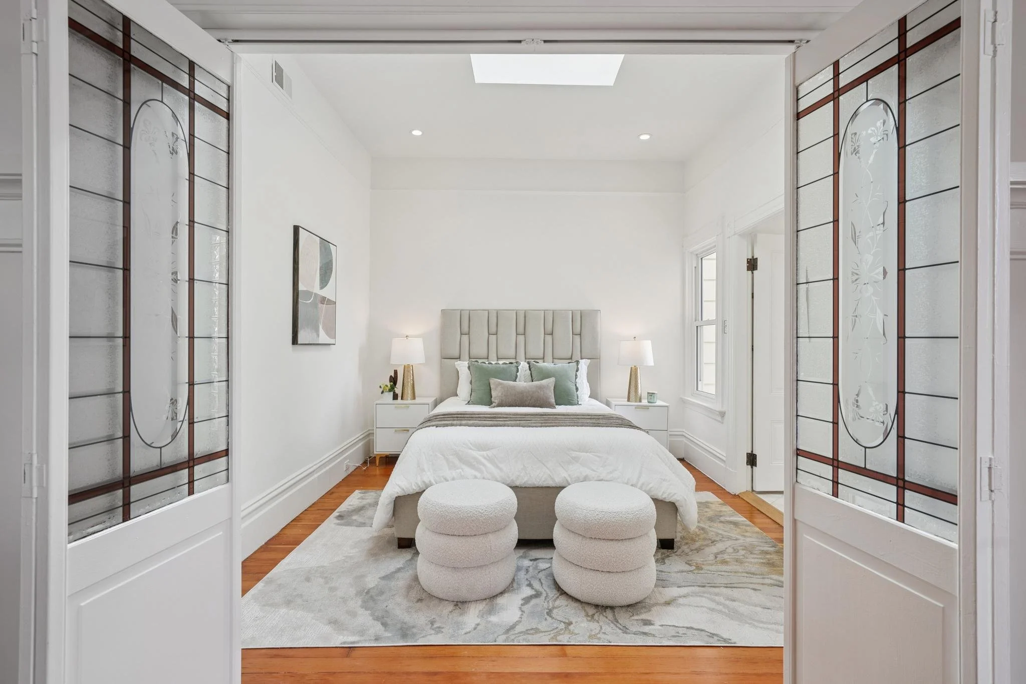 Modern bedroom with white walls, hardwood floors, a bed with a beige upholstered headboard, white bedding, decorative pillows, two white nightstands with lamps, two round white ottomans, and side windows. French doors with stained glass panels open i