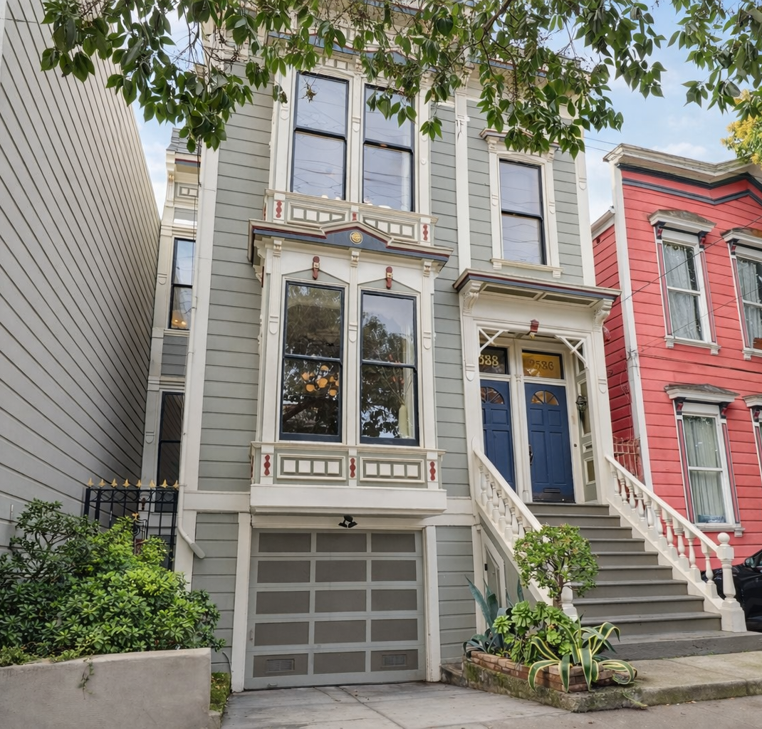 Three-story Victorian house painted light gray with decorative trim, tall windows, and a blue front door accessed by stairs, with a small garden and trees in front.