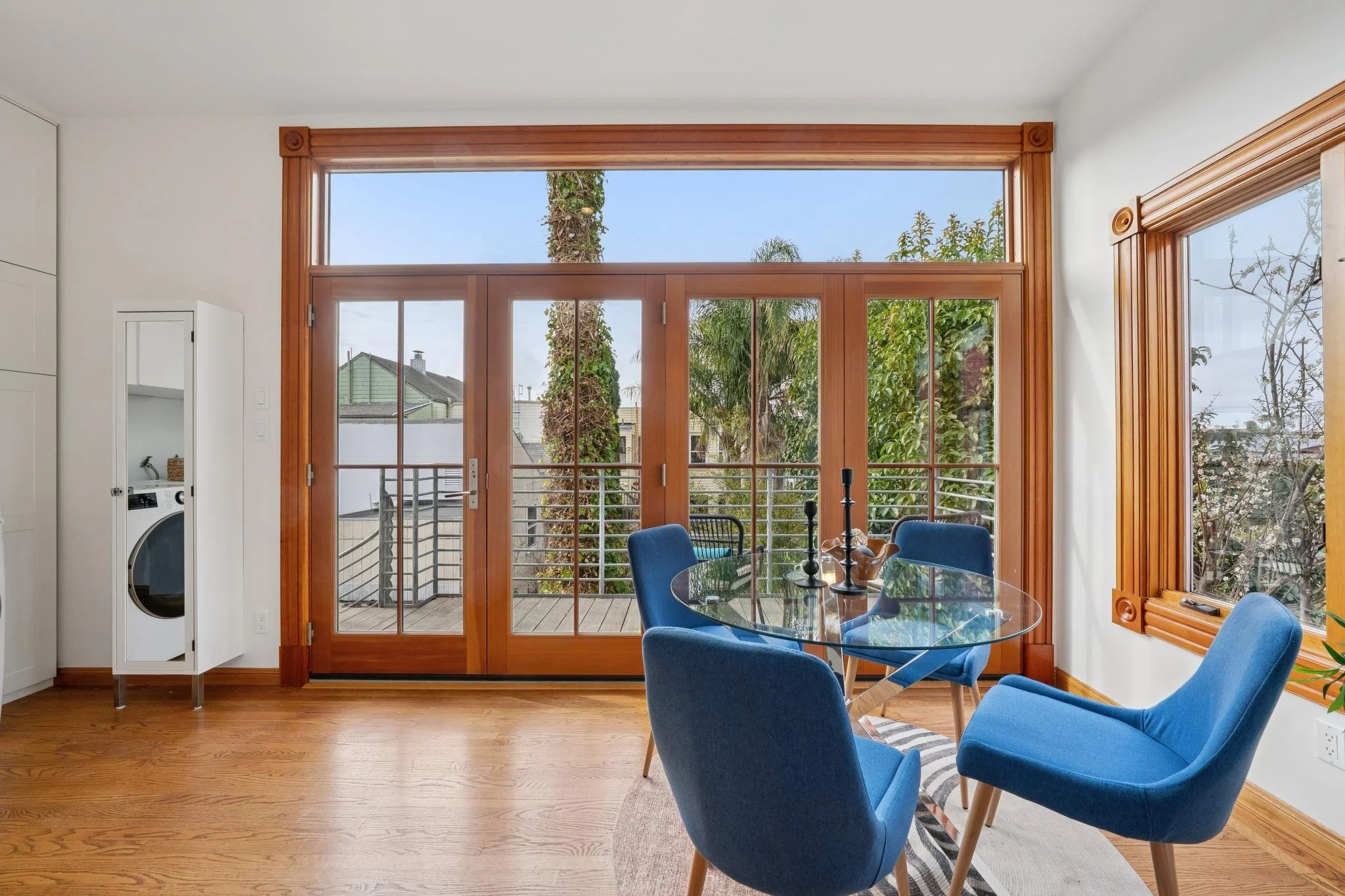 Interior view of a dining area with a glass table and four blue chairs, large wooden-framed glass doors leading to an outdoor balcony with trees and neighboring houses visible, and a corner with a laundry cabinet.