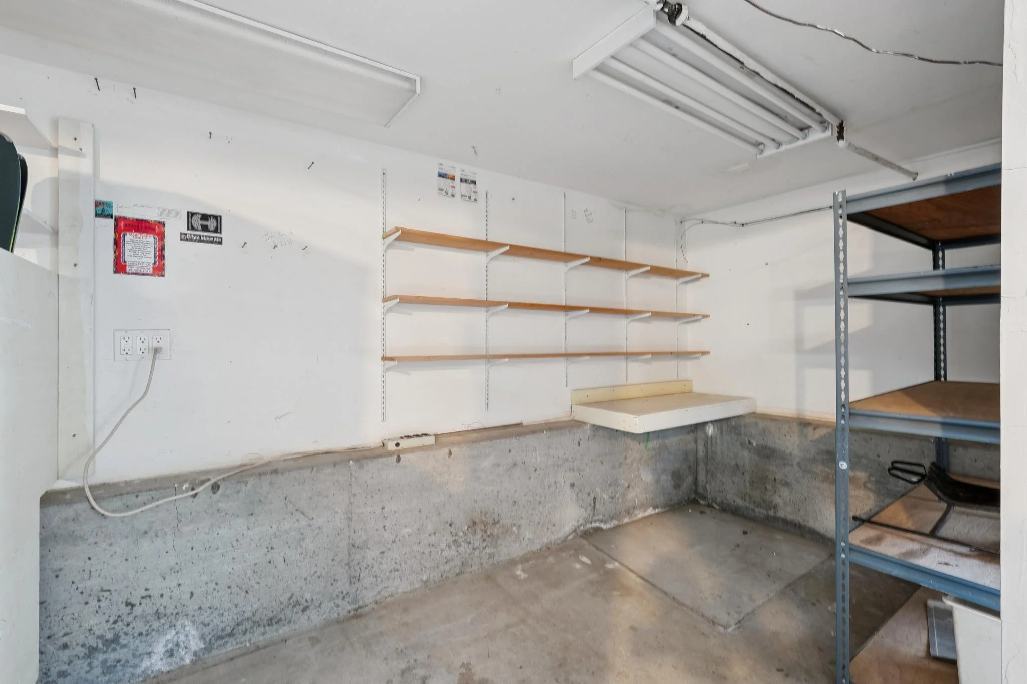 Empty garage with concrete floor, white walls, several orange-colored wooden shelves, metal shelving unit, and a power outlet.