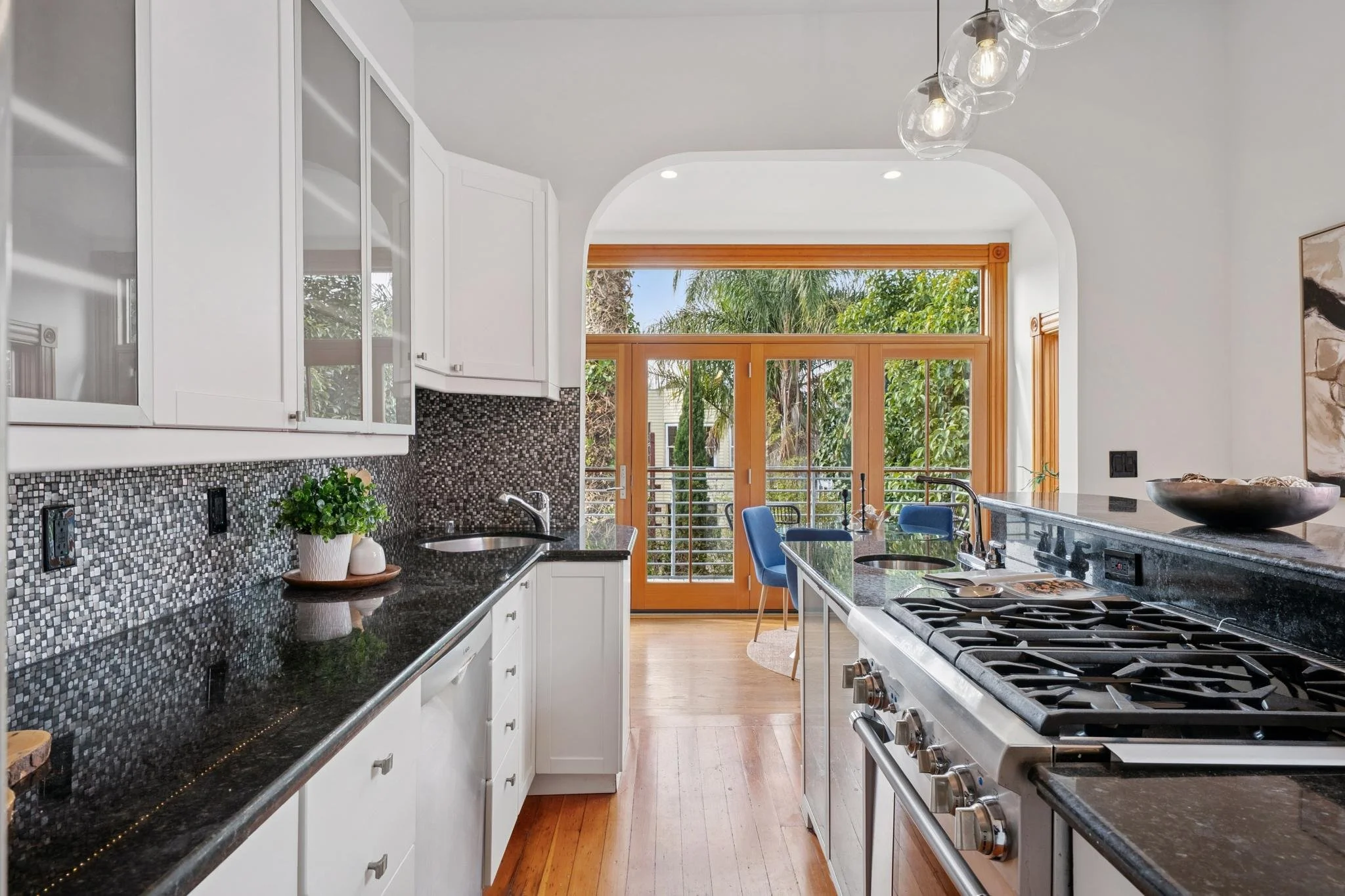 Modern kitchen with white cabinets, black granite countertops, mosaic backsplash, and a view of a dining area with blue chairs and large windows showing greenery outside.