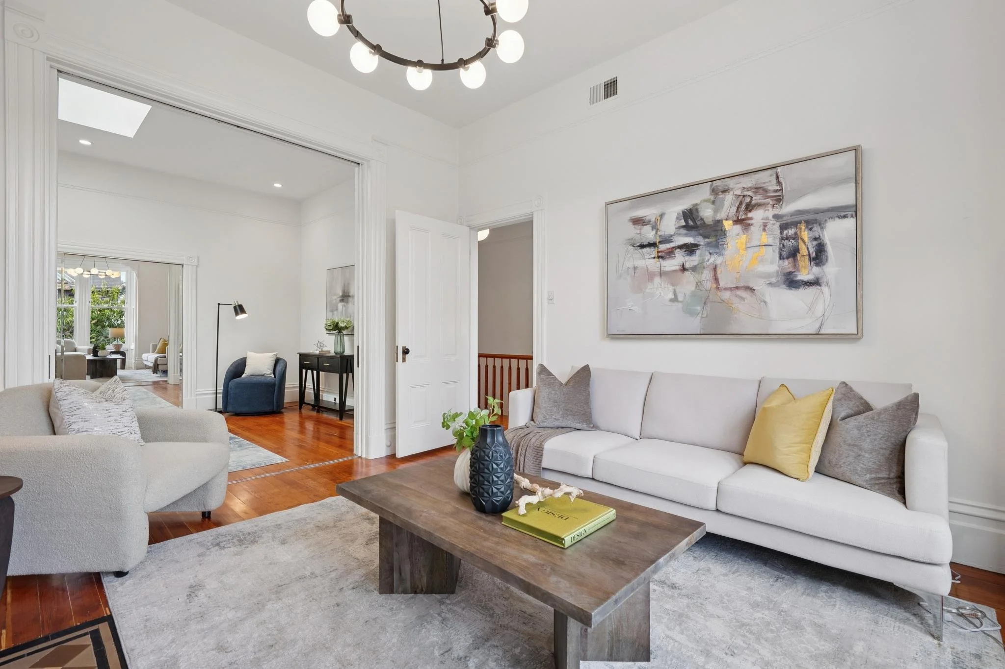 Living room with white walls and a white sofa with gray and yellow pillows, a wooden coffee table with decorations, and a large abstract painting above the sofa. Additional rooms are visible through open doorways.