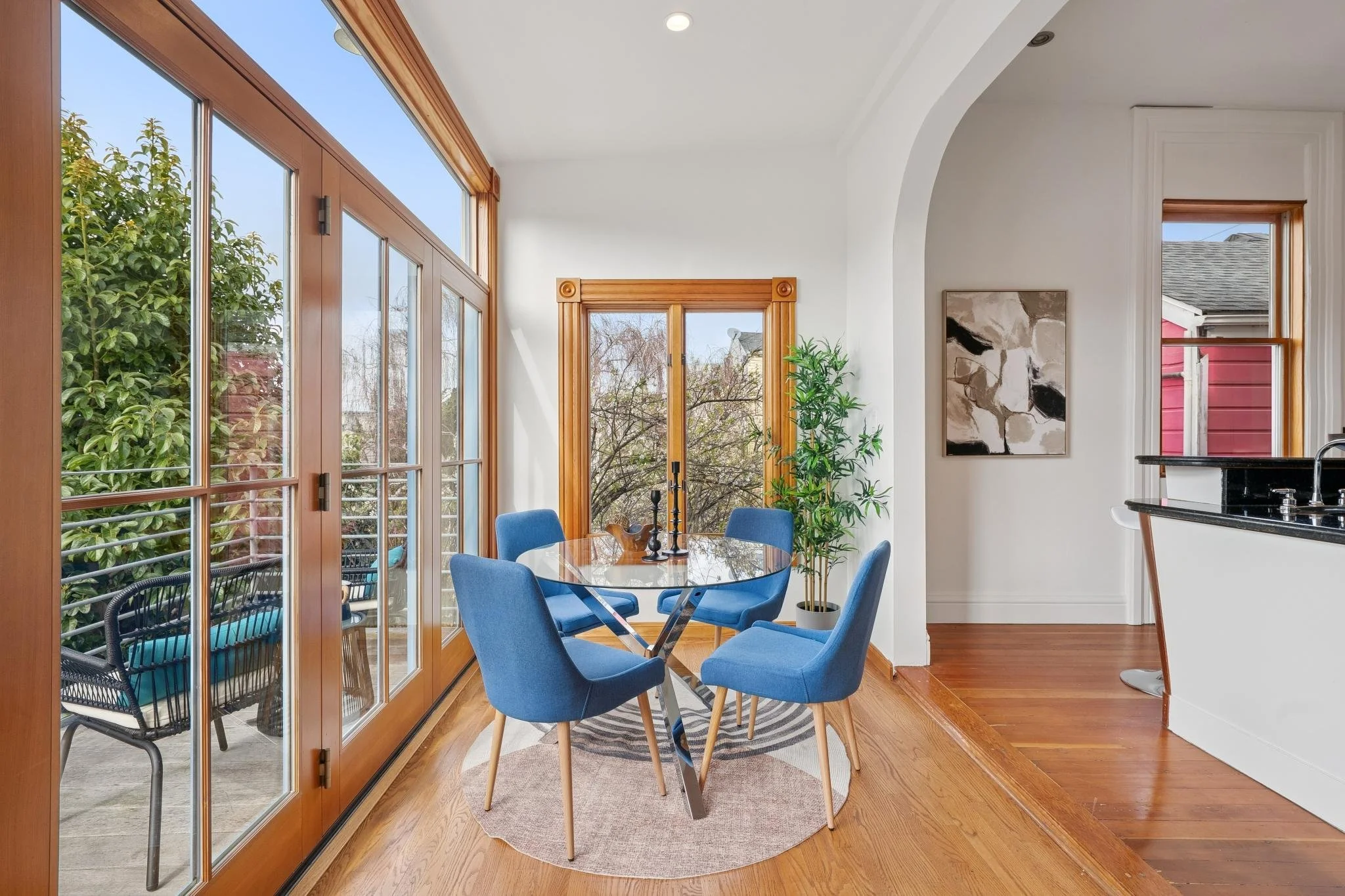 Dining area with a round glass table surrounded by five blue upholstered chairs, large windows and a sliding glass door with wooden frames, a potted plant, and view of outdoor balcony with chairs.