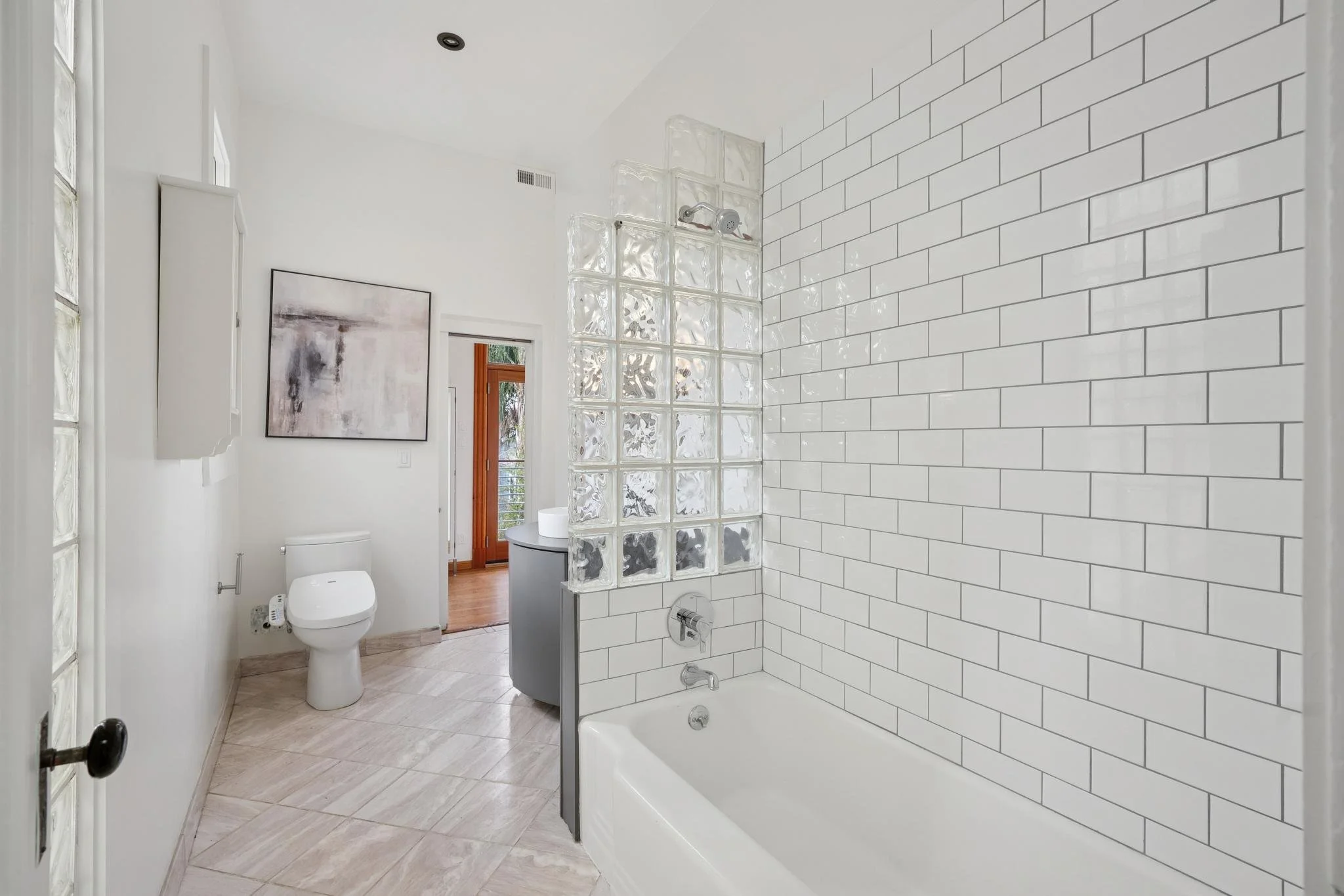 Bathroom with white subway tiles on the wall, a bathtub, a glass block wall, a toilet, and a small vanity sink. There is a window and a piece of wall art.