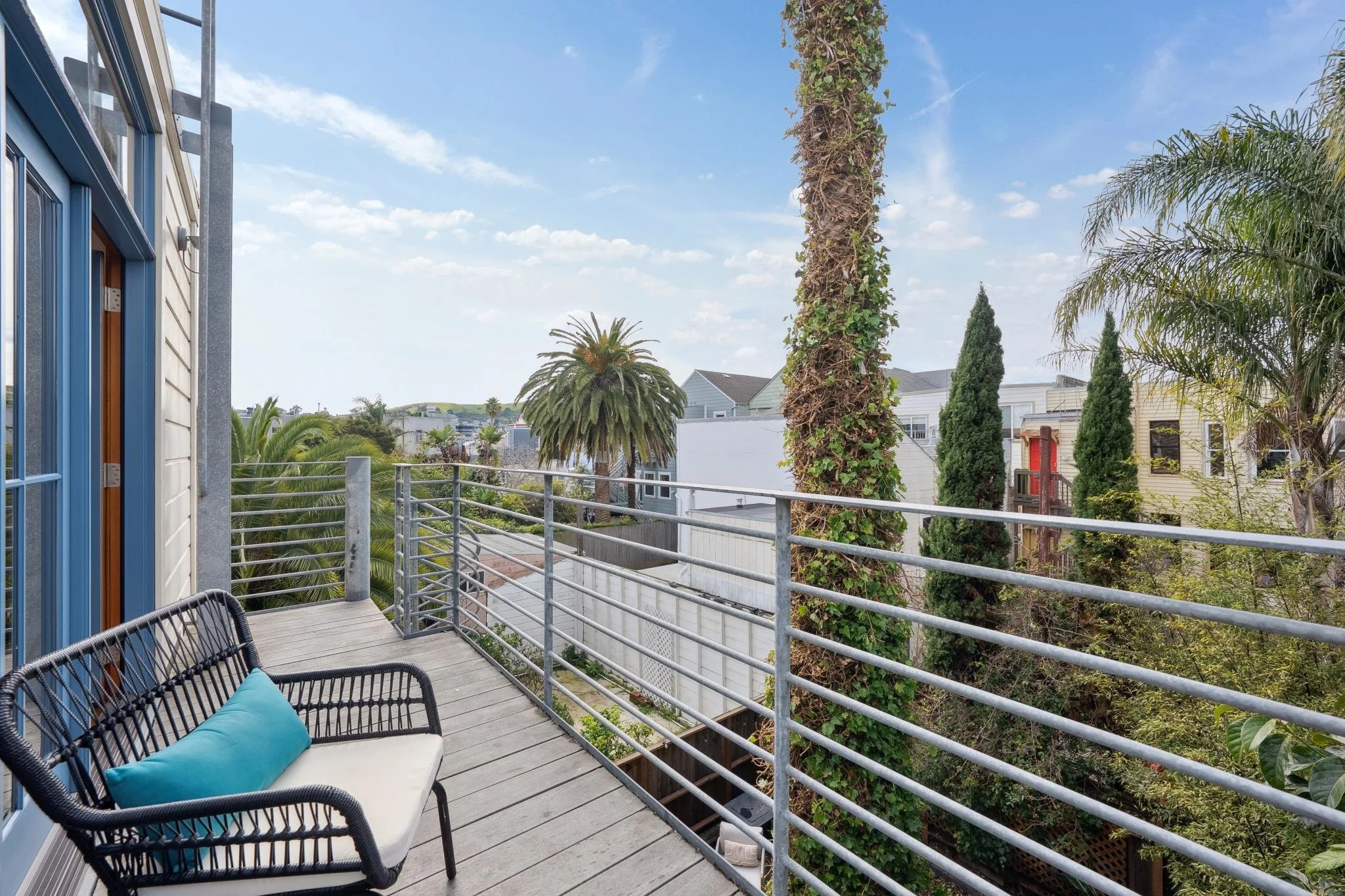 Balcony with black wicker chair and turquoise pillow, overlooking a garden with palm trees and houses under a partly cloudy sky.