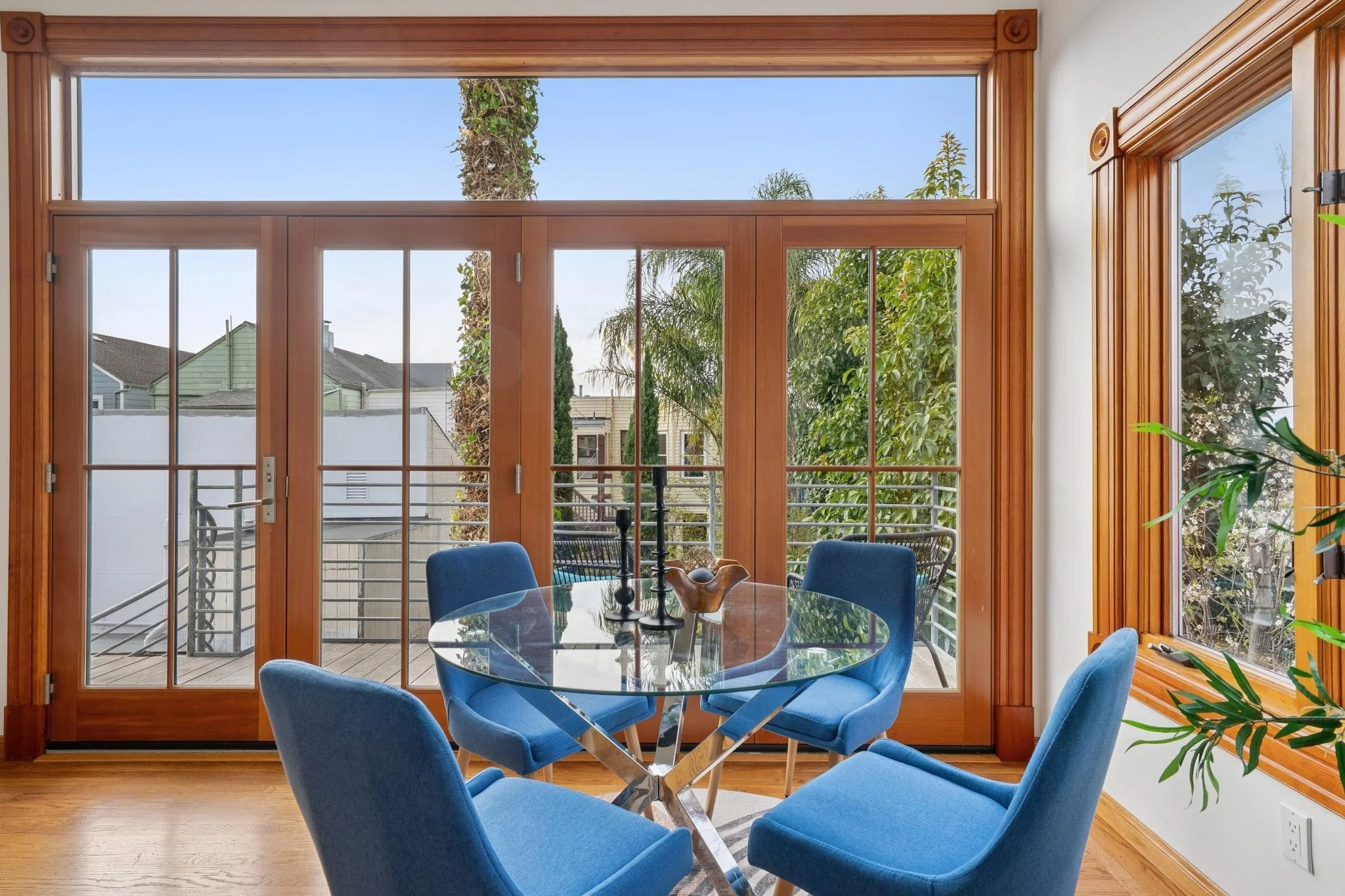 Dining room with a round glass table, six blue upholstered chairs, large wooden-framed glass doors leading to an outdoor balcony, and a window with similar framing showing green trees outside.