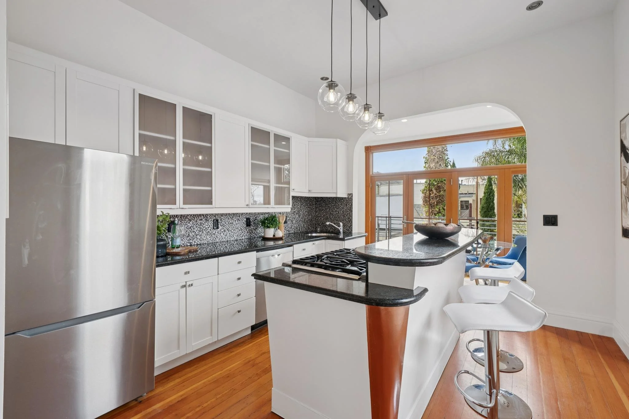 Modern white kitchen with black countertops and a small island, stainless steel refrigerator, glass-front cabinets, pendant lighting, and a large window with a view of trees outside.