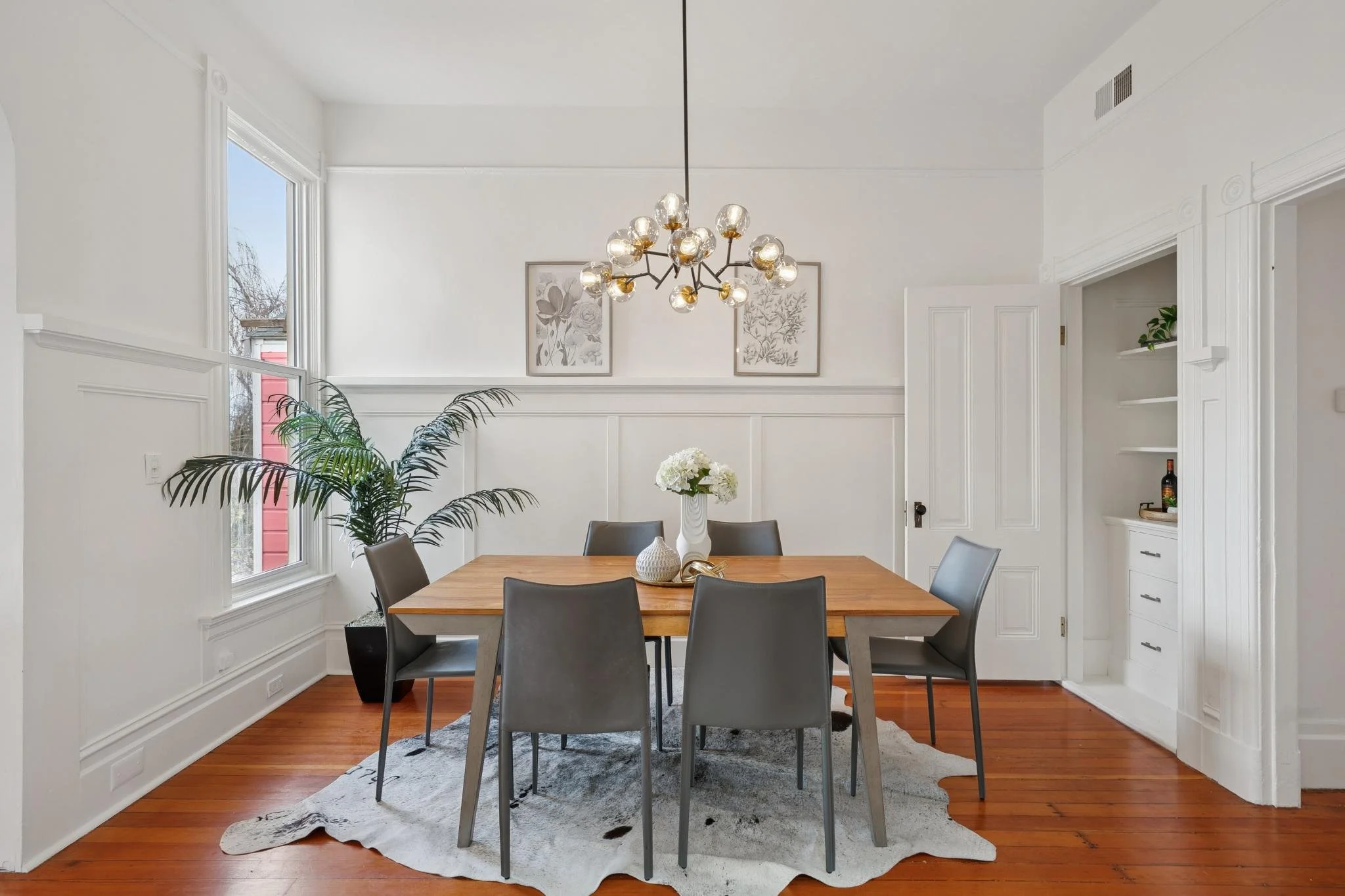 Dining room with white walls, a wooden table surrounded by six grey chairs, a large window, a black planter with a green plant, a modern chandelier, and framed botanical artwork on the wall.