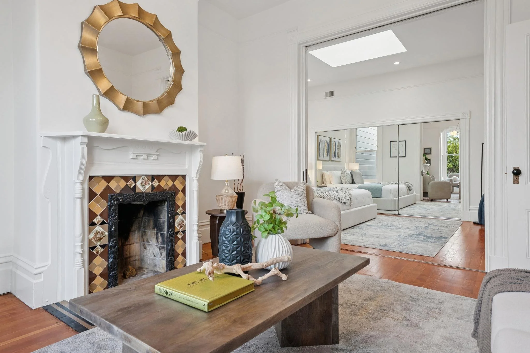 Living room with white walls, a fireplace with decorative tiles, a wooden coffee table with books and decorative objects, beige sofas, and a mirrored sliding door leading to another sitting area with armchairs and a bright window.
