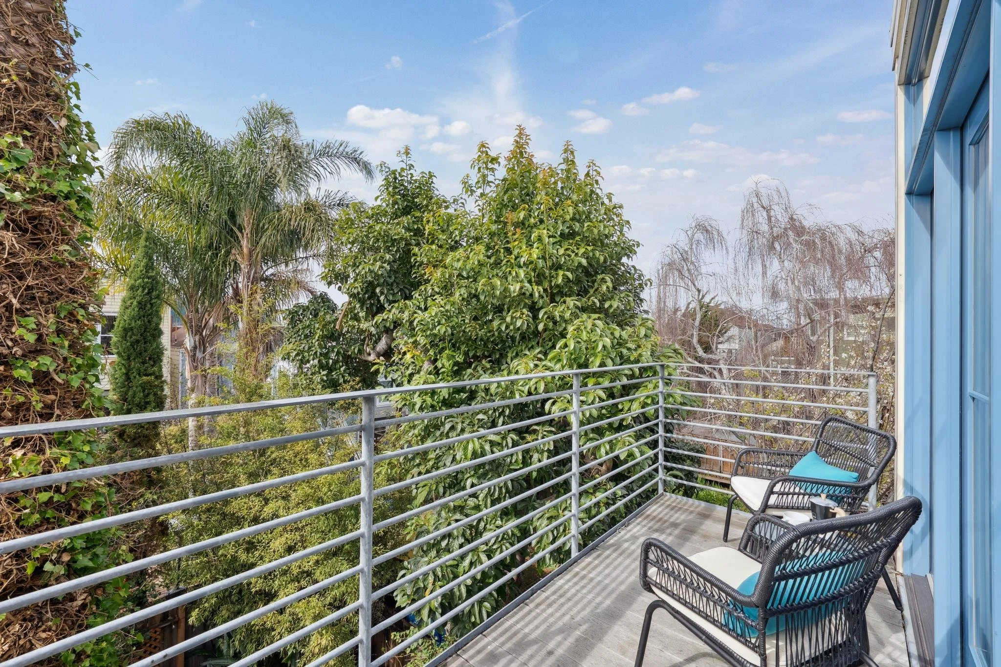 Balcony with two black wicker chairs with white cushions and blue pillows, overlooking green trees, palm, and a blue sky.