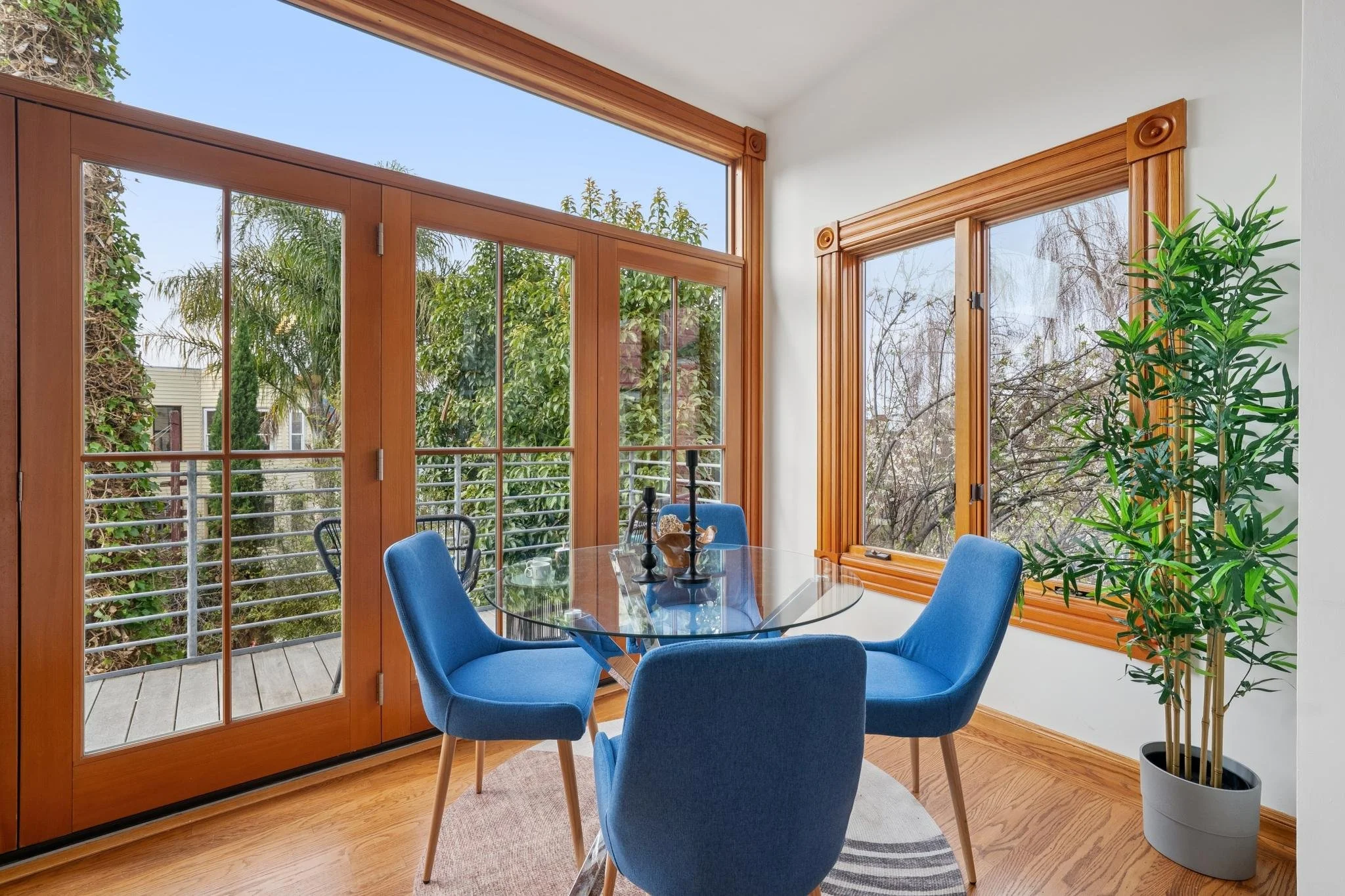 A bright dining area with large wooden framed glass doors and windows, a round glass table, four blue chairs, a tall green plant, and a view of trees outside.