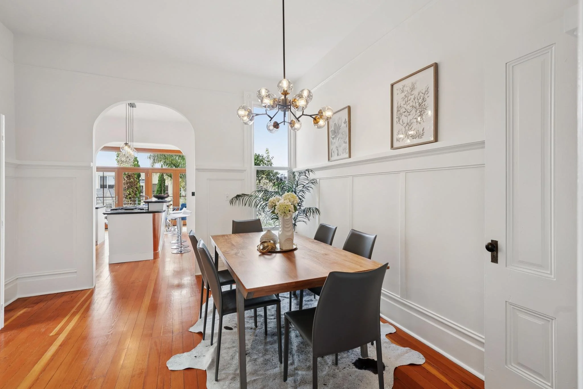 Dining room with wooden table, black chairs, and chandelier, next to a white wainscoted wall with framed artwork. Open archway leads to kitchen with barstools and sliding glass doors to outdoor space.