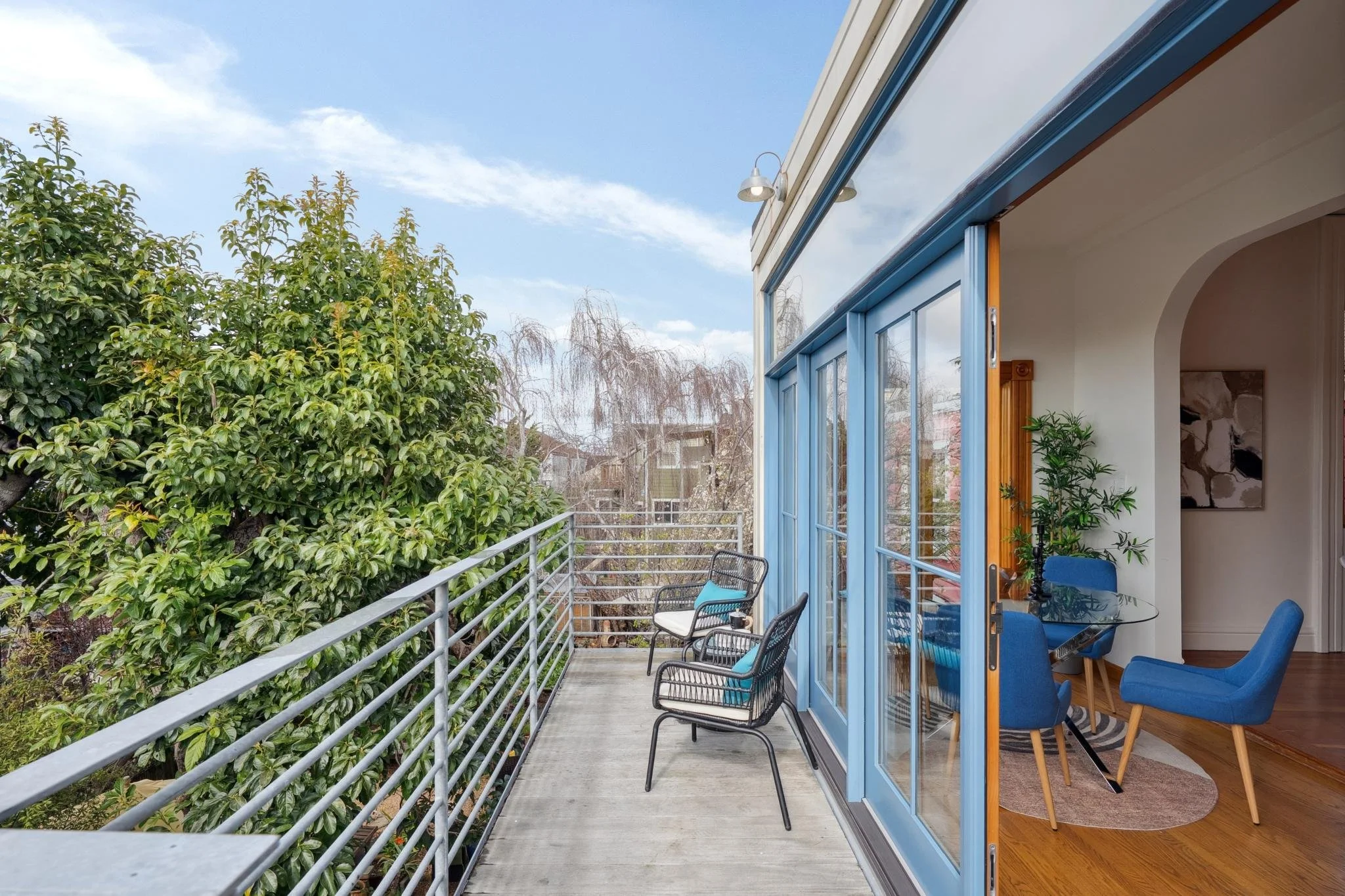 View of a balcony with chairs, overlooking a tree with bare branches, adjacent to a modern living room with blue chairs and a glass table.